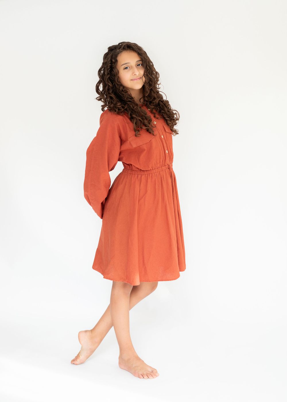Model wearing flowy rust-colored dress poses elegantly with curly hair against white backdrop.