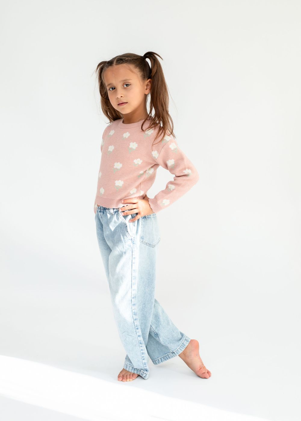 Young person in pink polka dot sweater and light jeans poses casually against white background.