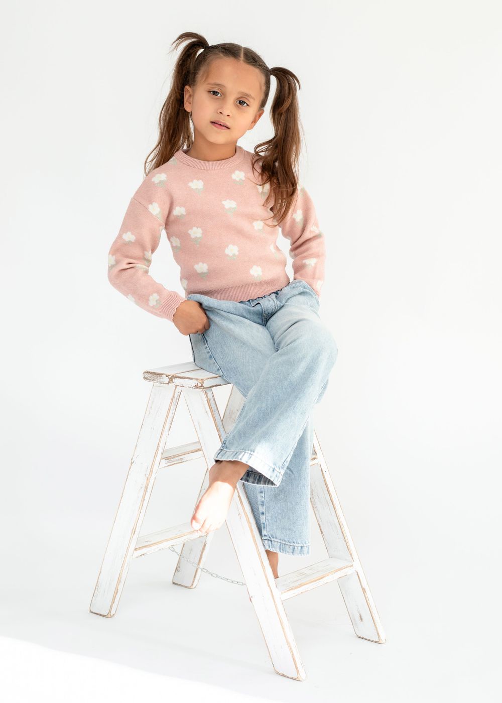 Model in pink polka dot sweater sits on white ladder wearing light wash jeans.