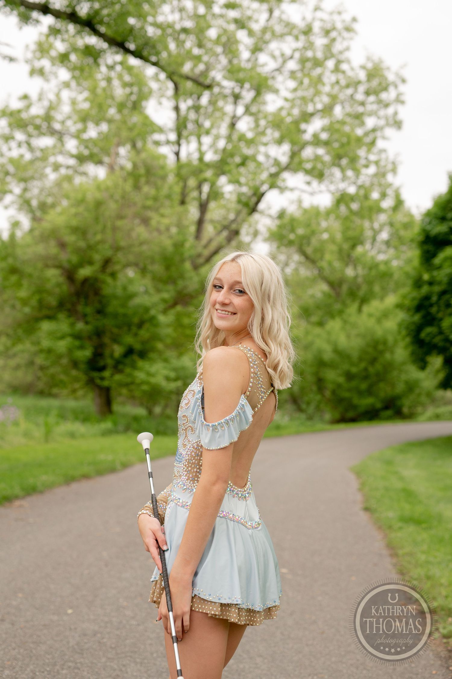 Pageant winner in a light blue dress holds a scepter while posing on a garden path.
