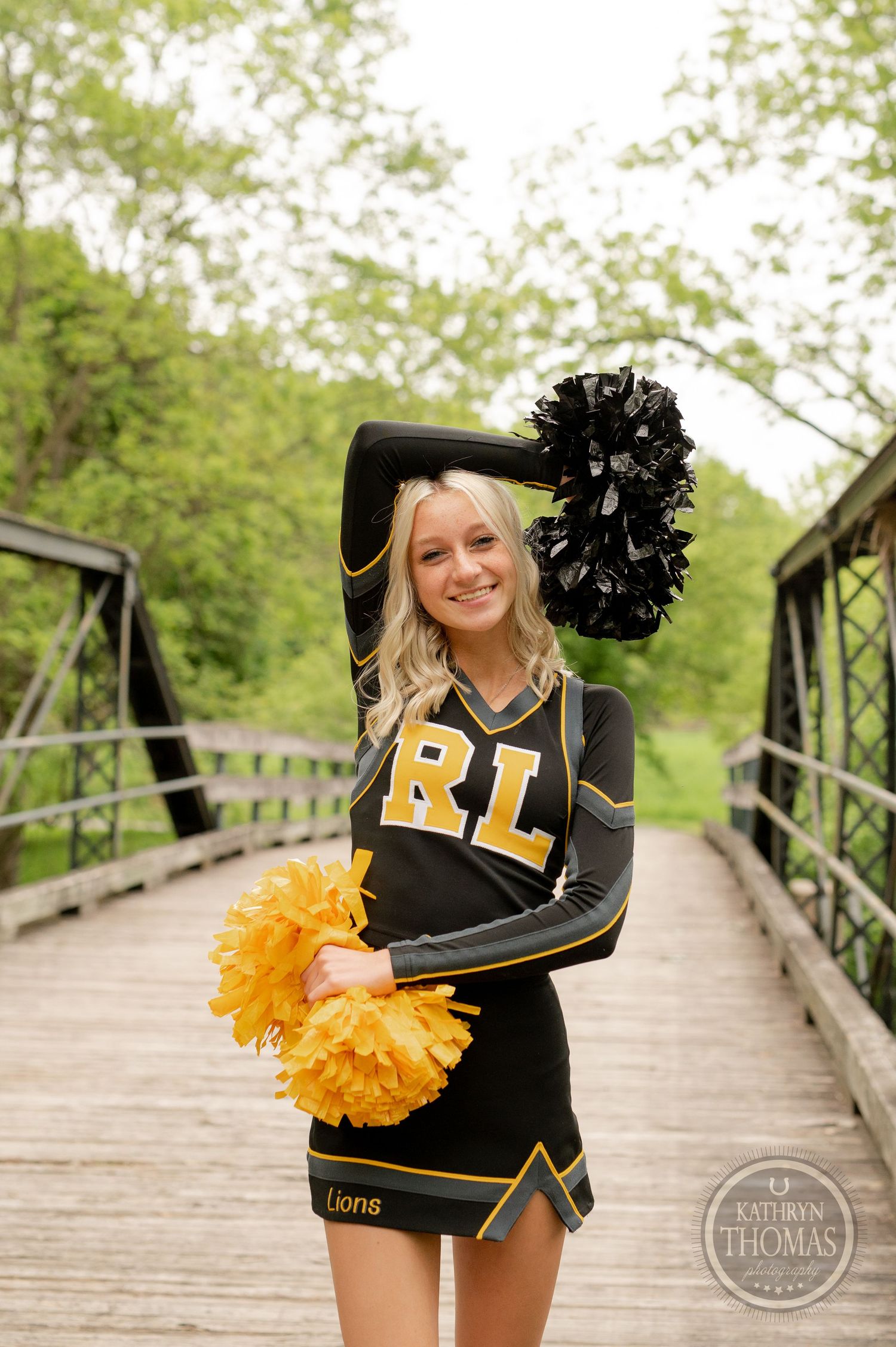 Cheerleader in black uniform with gold letters poses with pom poms on wooden bridge in park setting.