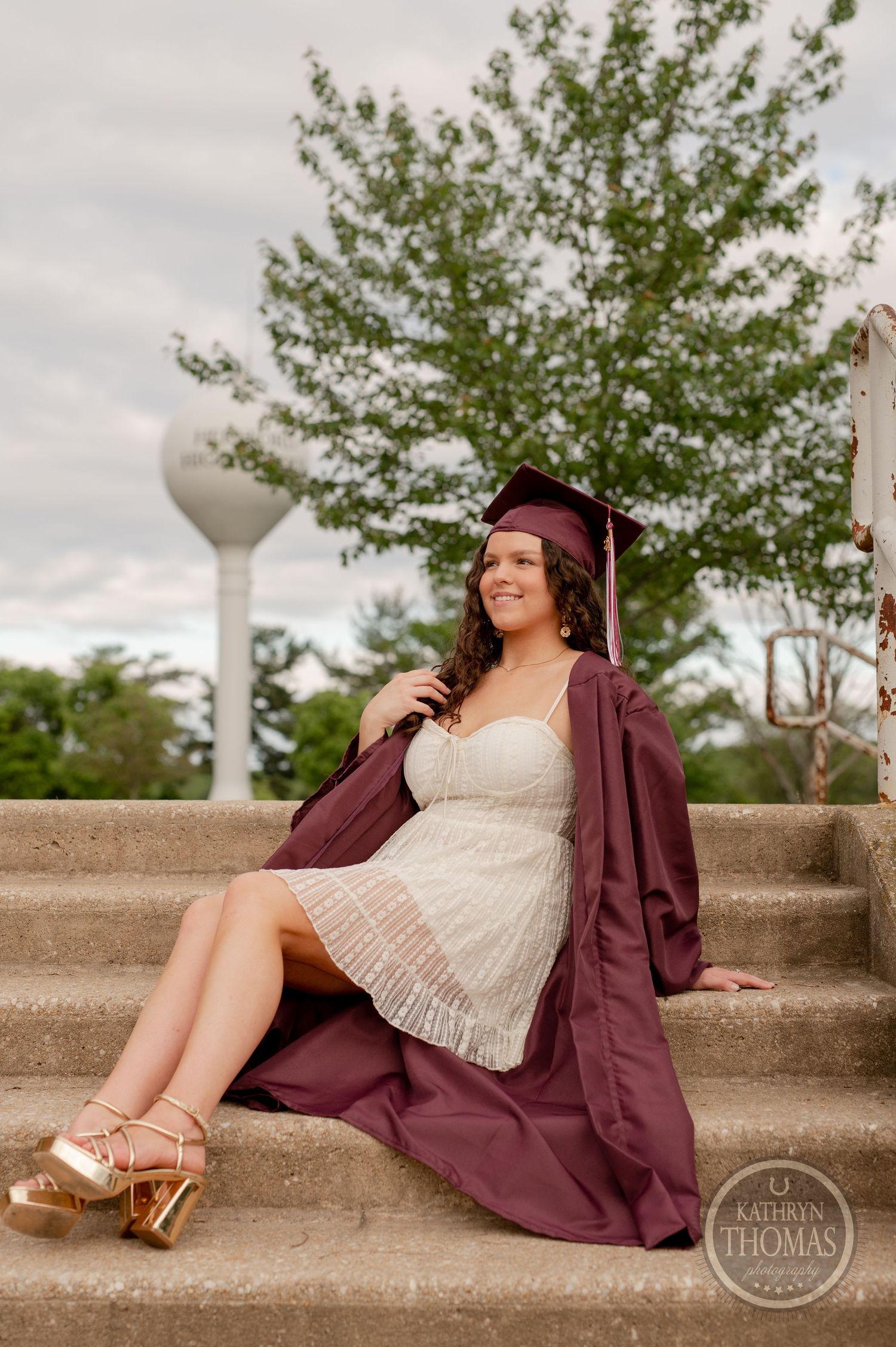 Graduate in maroon cap and gown poses elegantly on outdoor concrete steps.