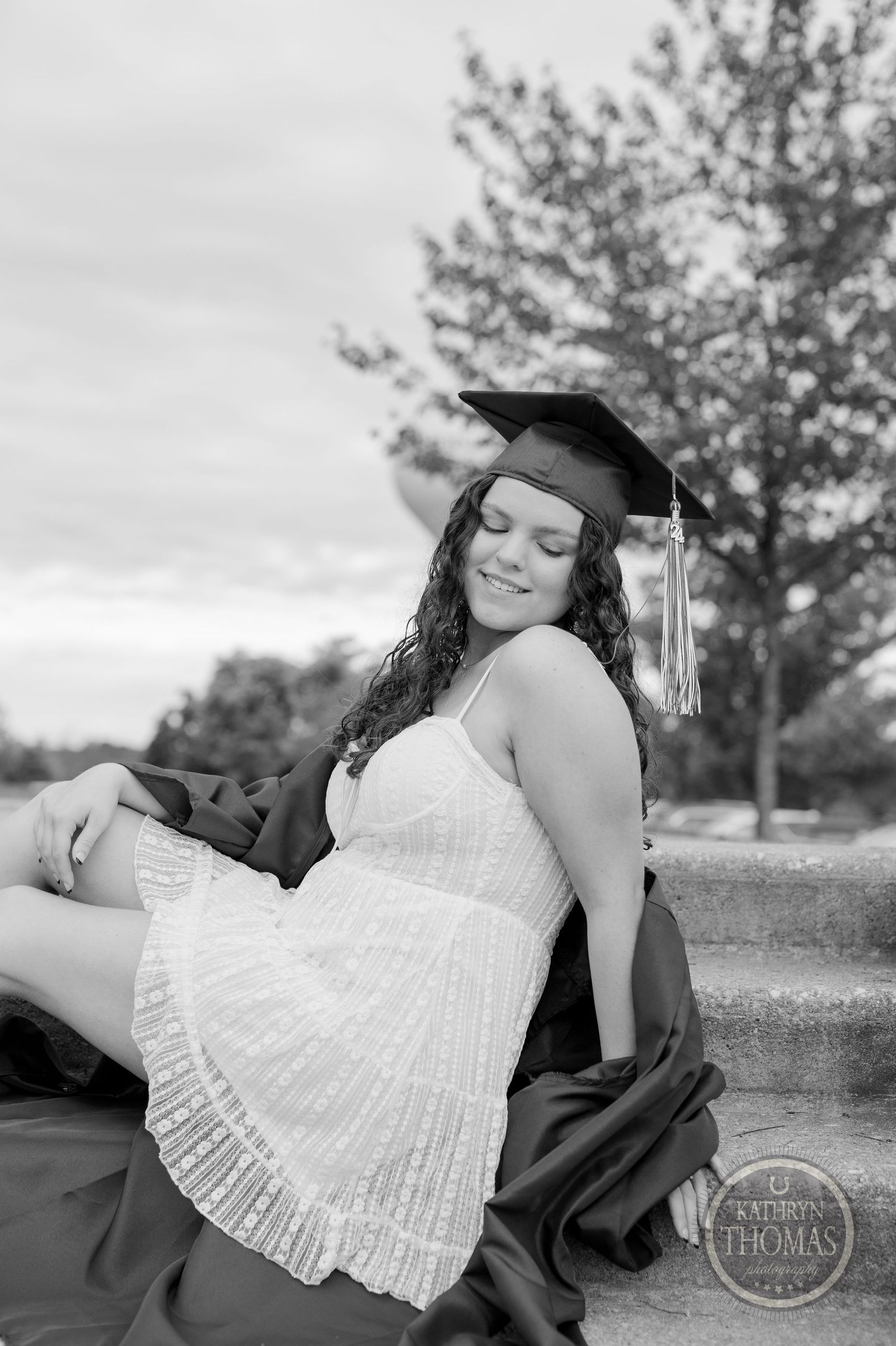 Black and white portrait of graduate in cap and white dress sitting on steps.