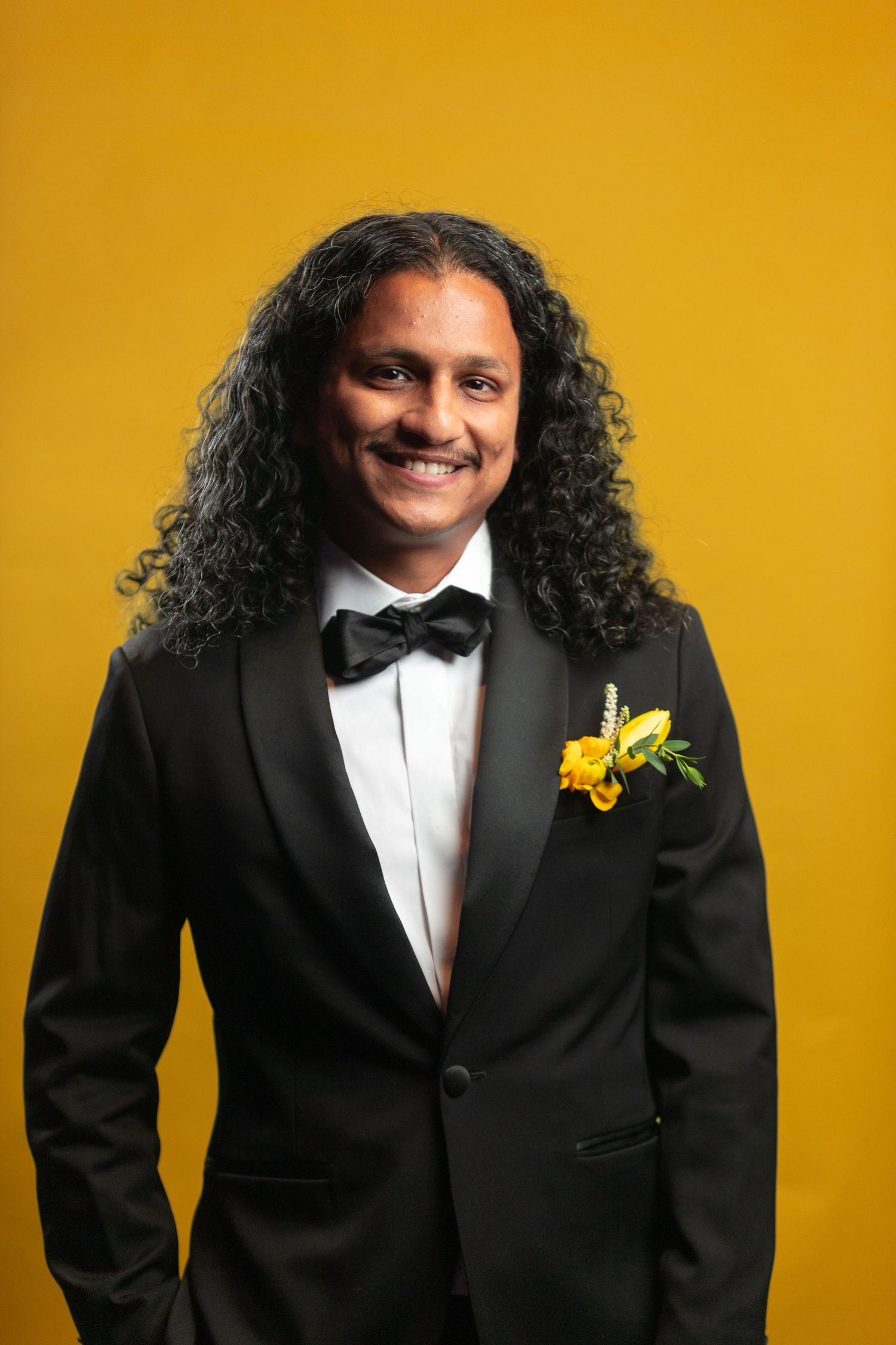 Person in tuxedo with yellow boutonniere smiling against yellow backdrop.