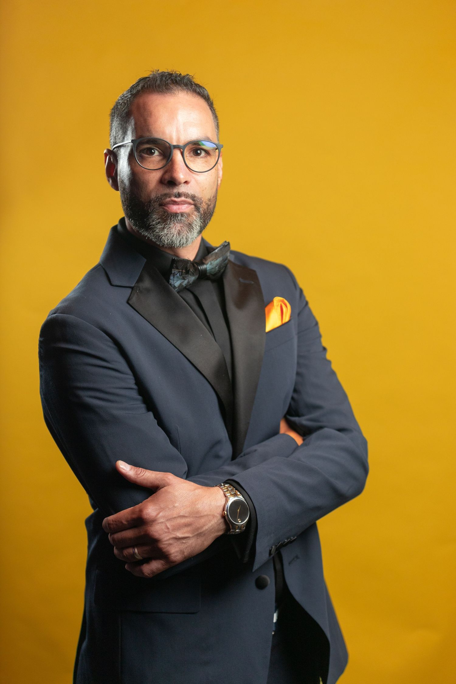 Professional portrait of a well-dressed man in a dark suit with glasses against a yellow backdrop.