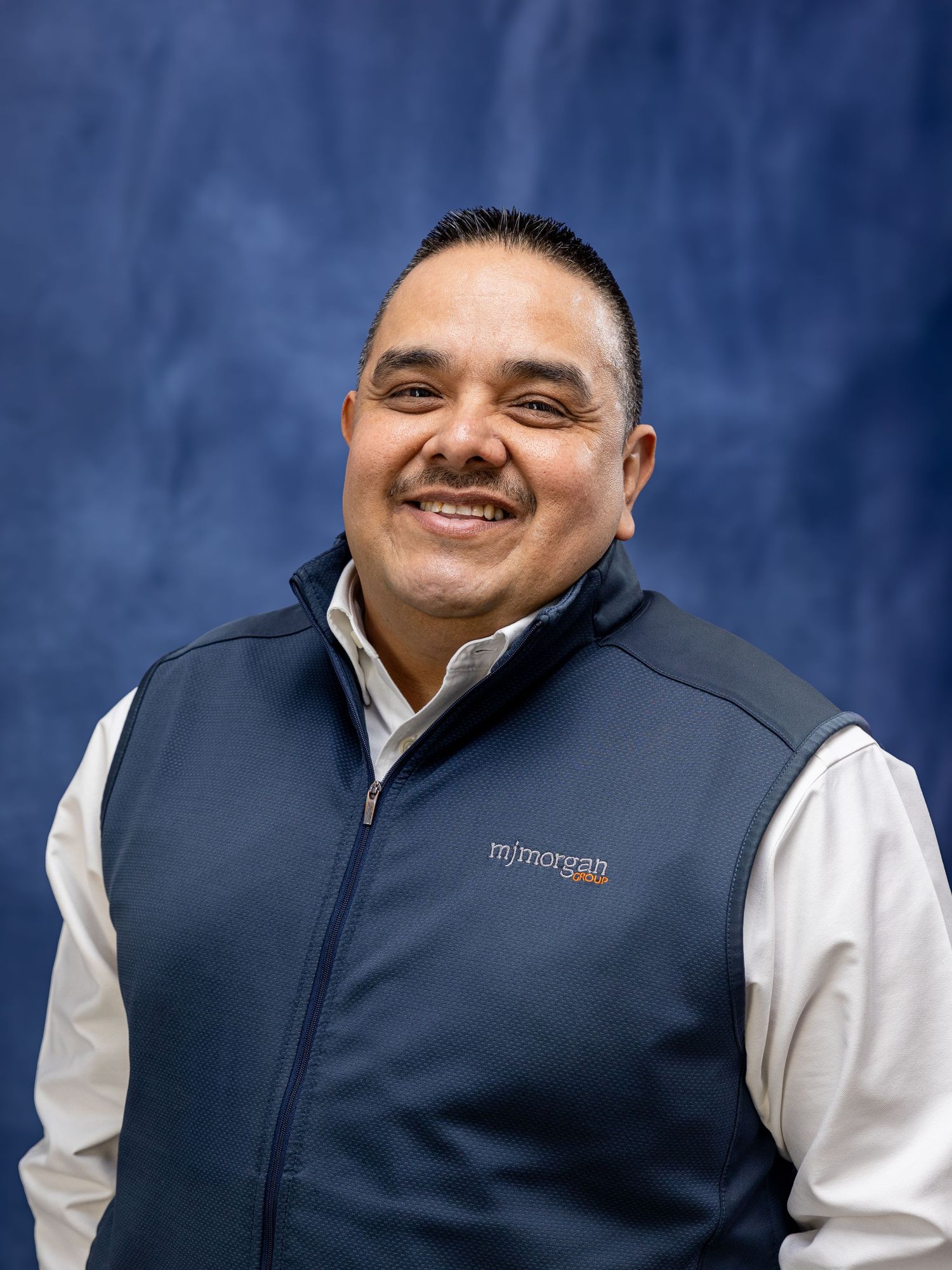 Professional headshot of a person in a navy blue vest and white dress shirt against a blue background.