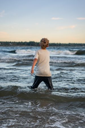 A child wading in the ocean waves during sunset with wet clothes.