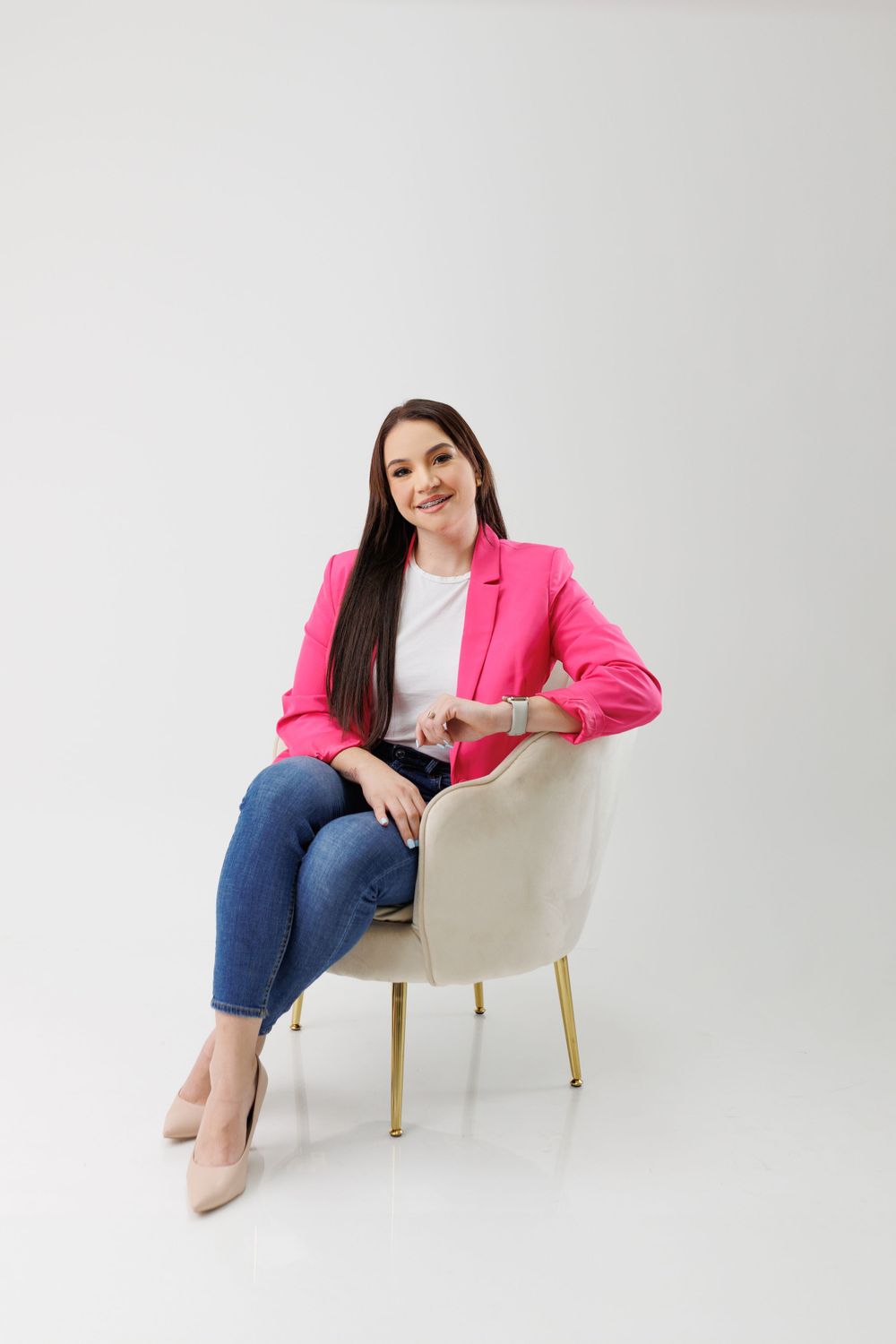 Model in pink blazer and jeans relaxing in white armchair during studio photoshoot.