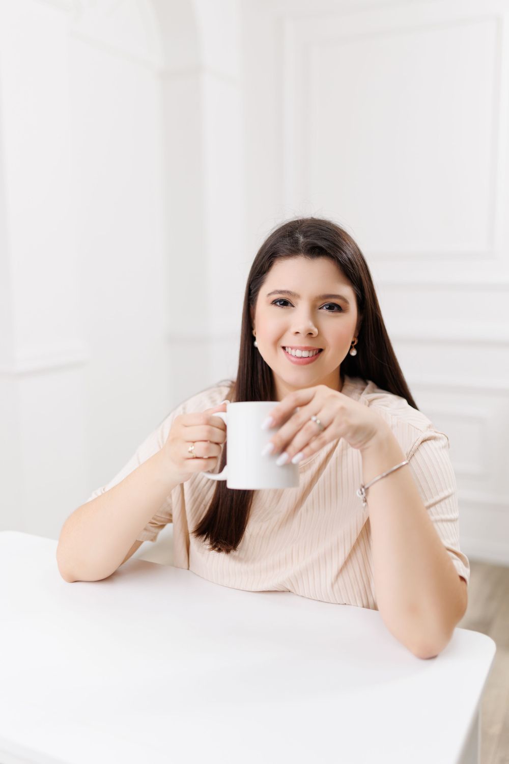 Woman in beige sweater holding white coffee cup while sitting at table.