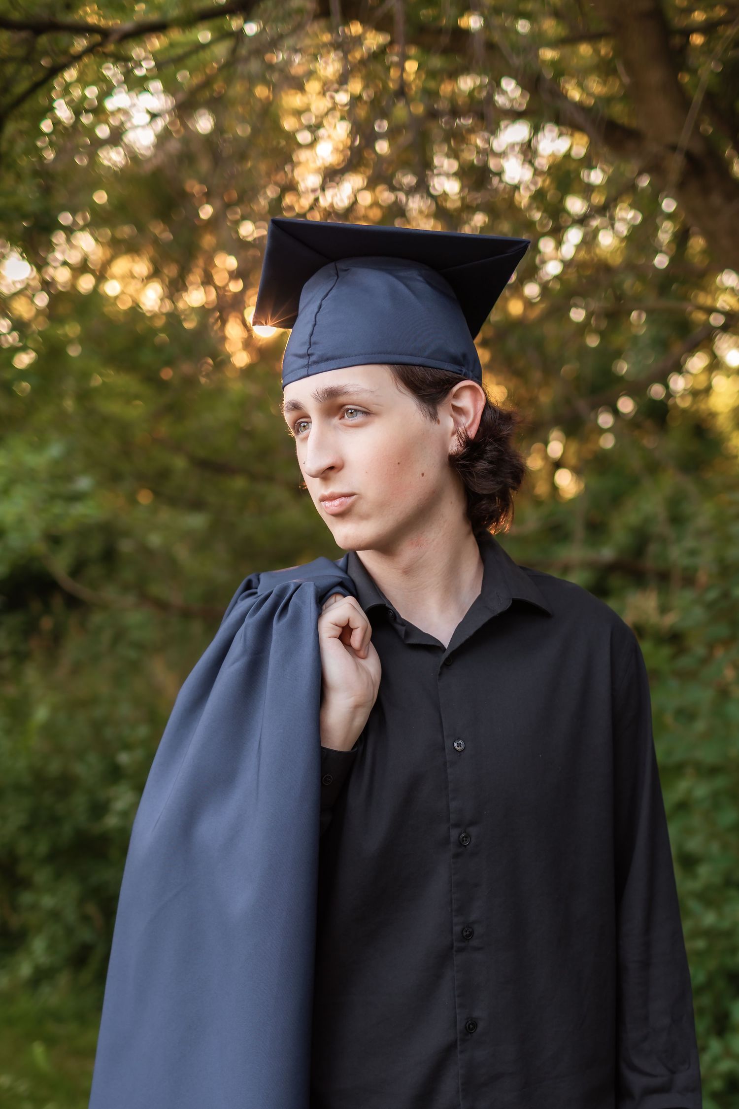 A graduate in a black cap and gown stands outdoors with golden sunlight filtering through trees in the background.