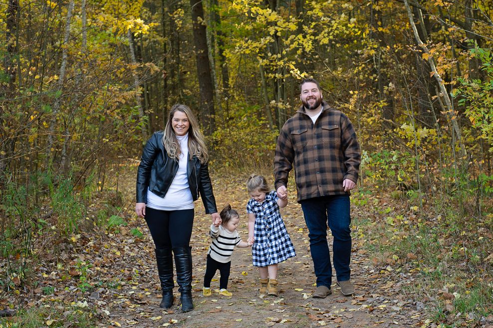 family of 4, parents and 2 young girls walk hand in hand on a forest trail in the fall with the bright changing leaves behind them