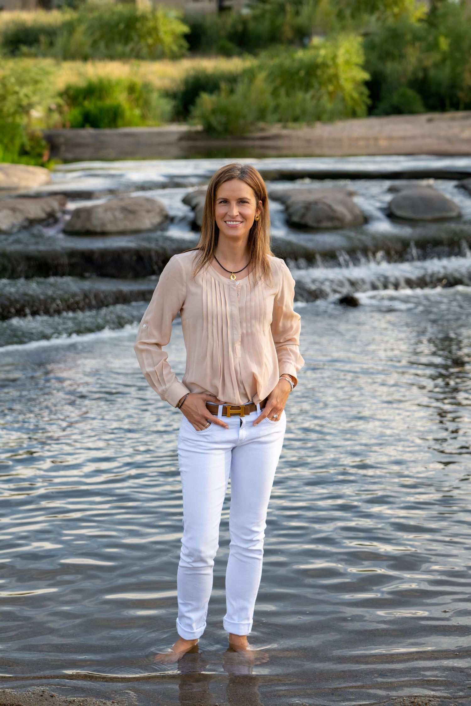 A woman stands ankle-deep in flowing river water wearing white pants and a peach colored blouse.