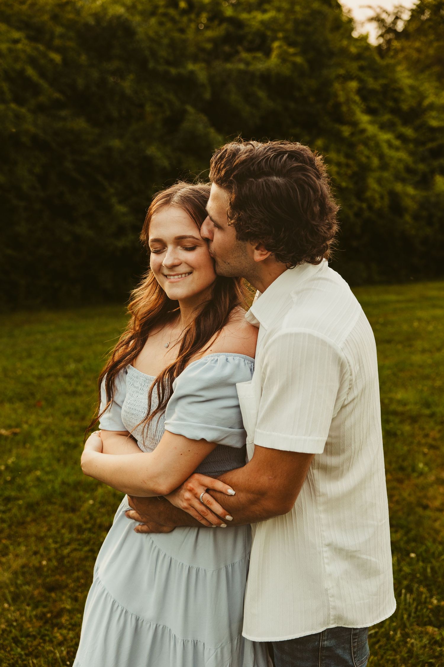 A tender embrace between a couple in a grassy field at golden hour, she in a blue dress and he in a white shirt.