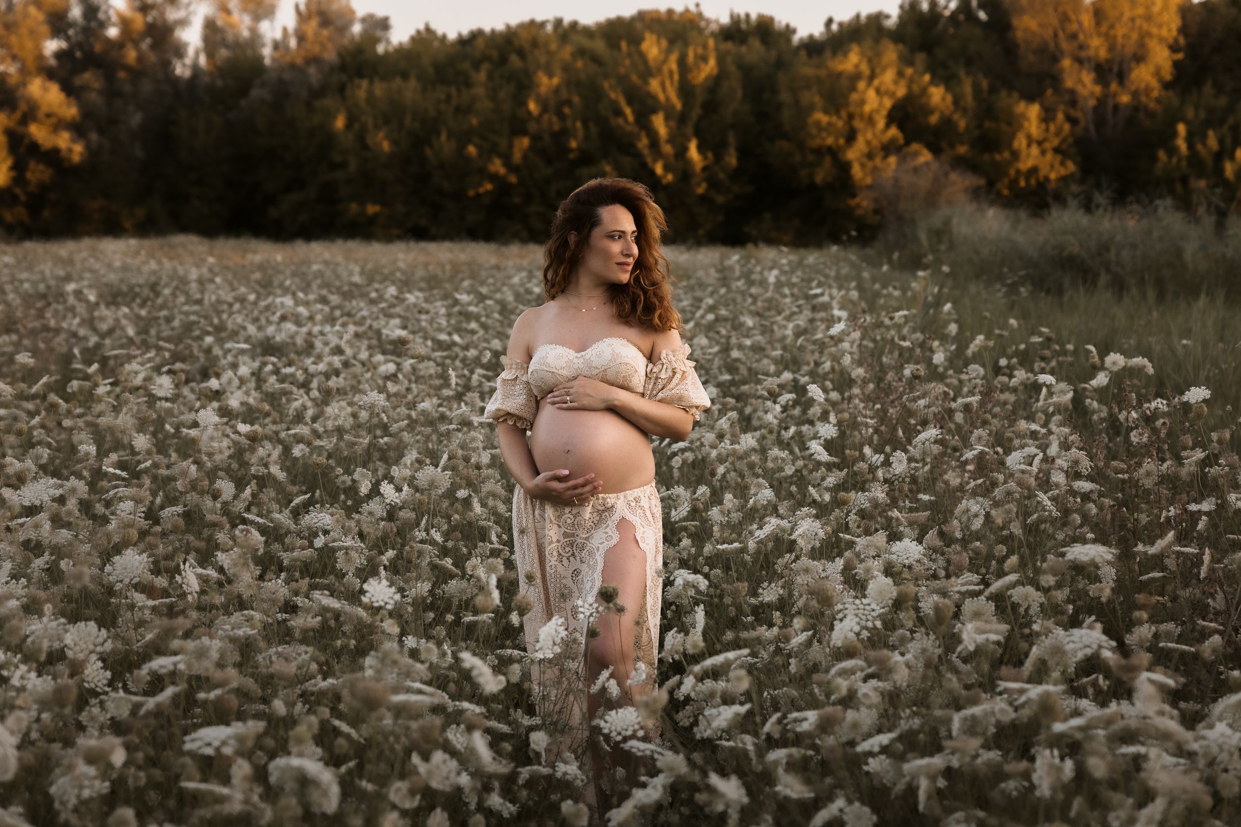 A dreamy maternity scene in a wildflower field at golden hour with warm autumn trees in the background.
