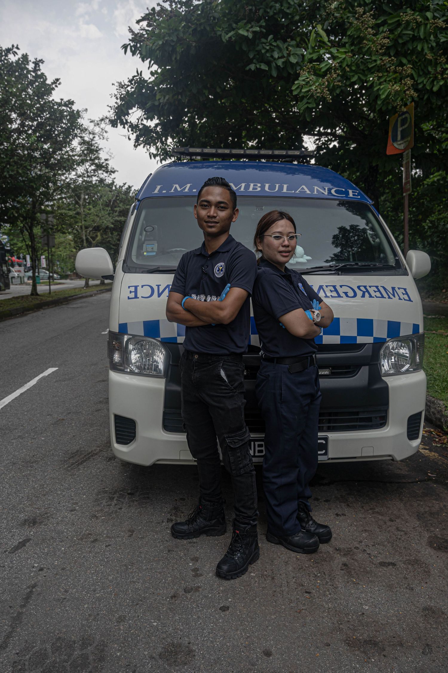 Two emergency medical professionals pose confidently in front of their ambulance vehicle on a tree-lined street.