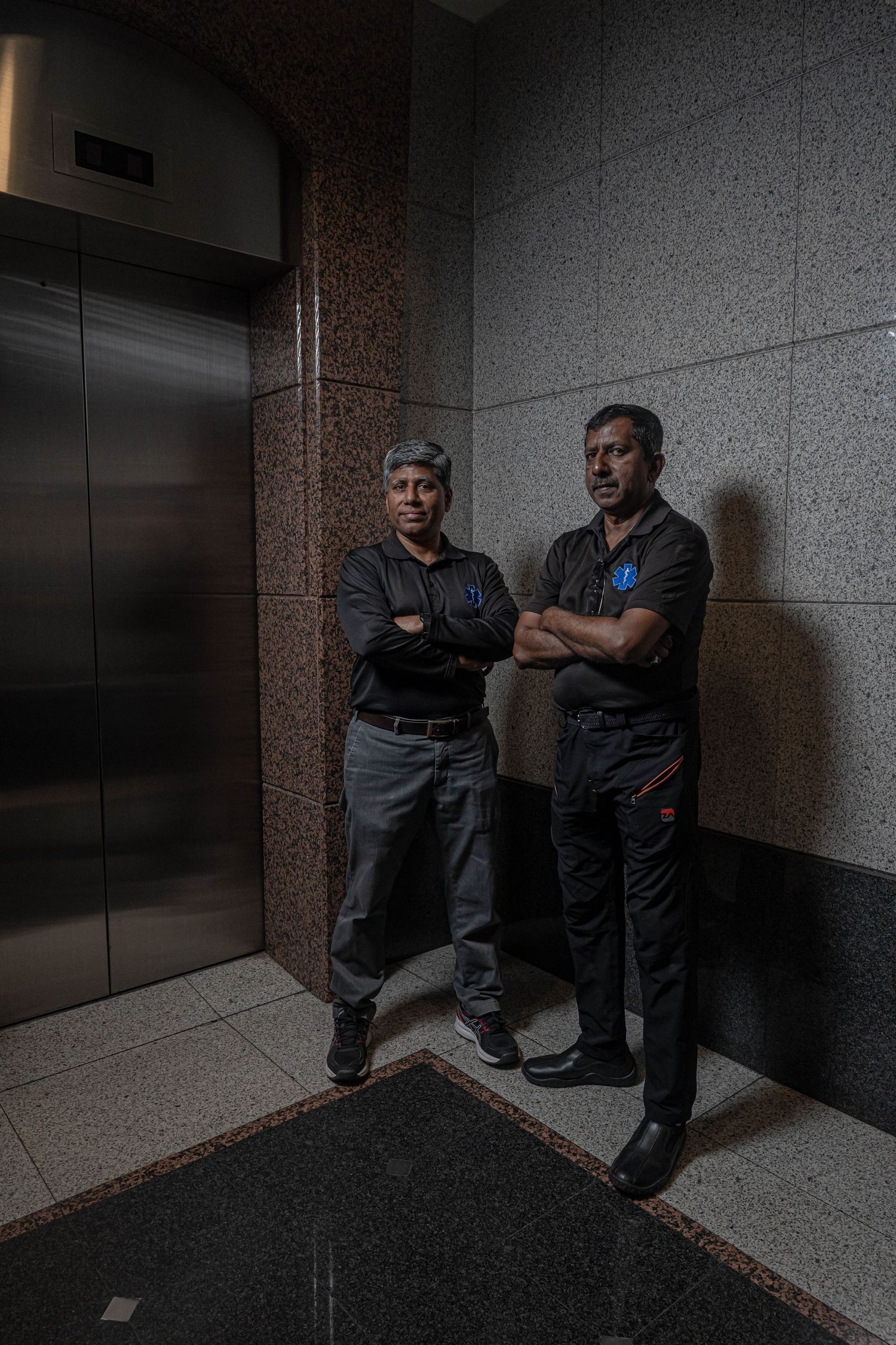 Two uniformed personnel stand with crossed arms near an elevator in a dimly lit hallway.