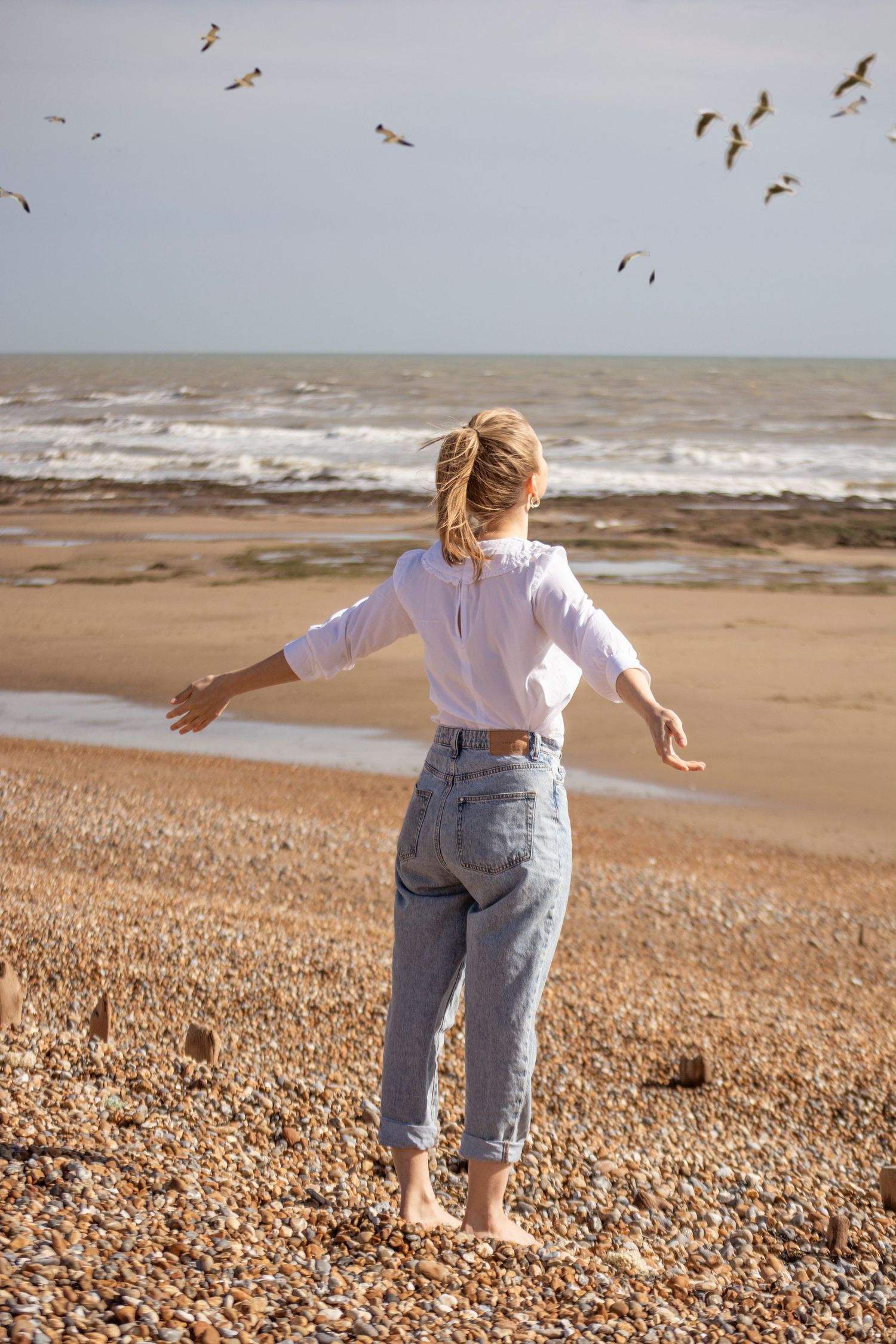 photograph of a female in blue jeans and a white top, arms spread wide, looking out at sea, seagulls ahead