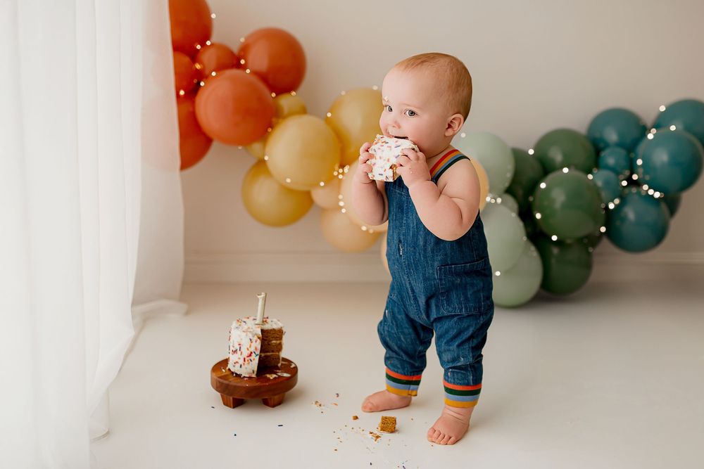 Baby boy in denim overalls enjoying cake smash session with rainbow balloon backdrop at Summerville SC studio