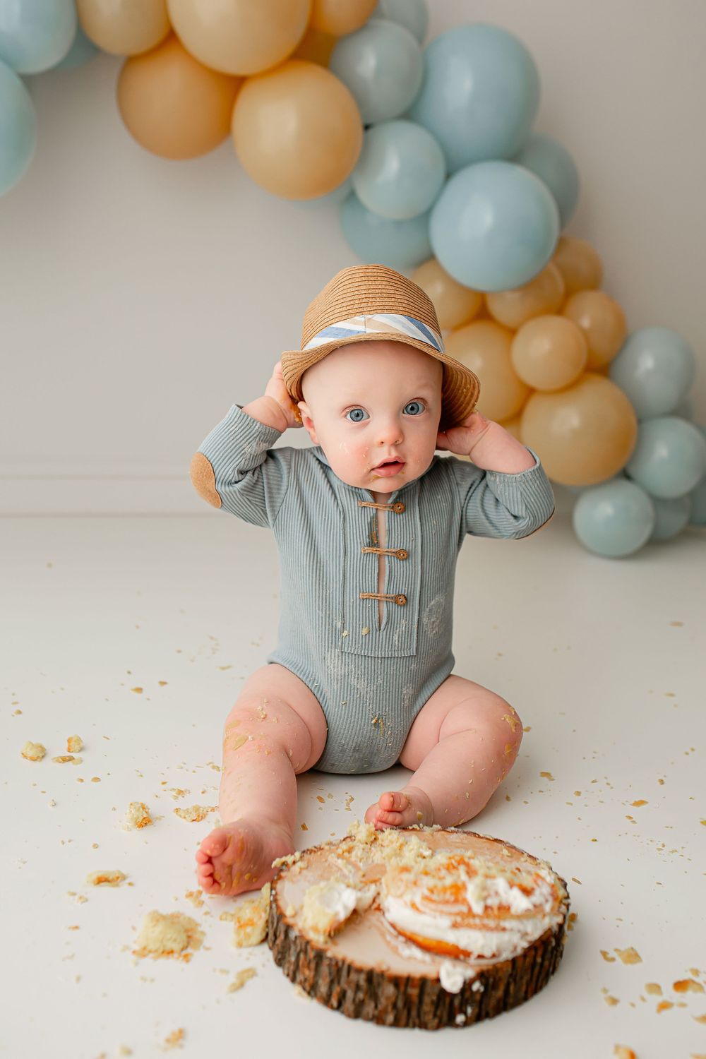 Baby wearing straw hat during cake smash session with sage and peach balloon arch at Summerville SC studio
