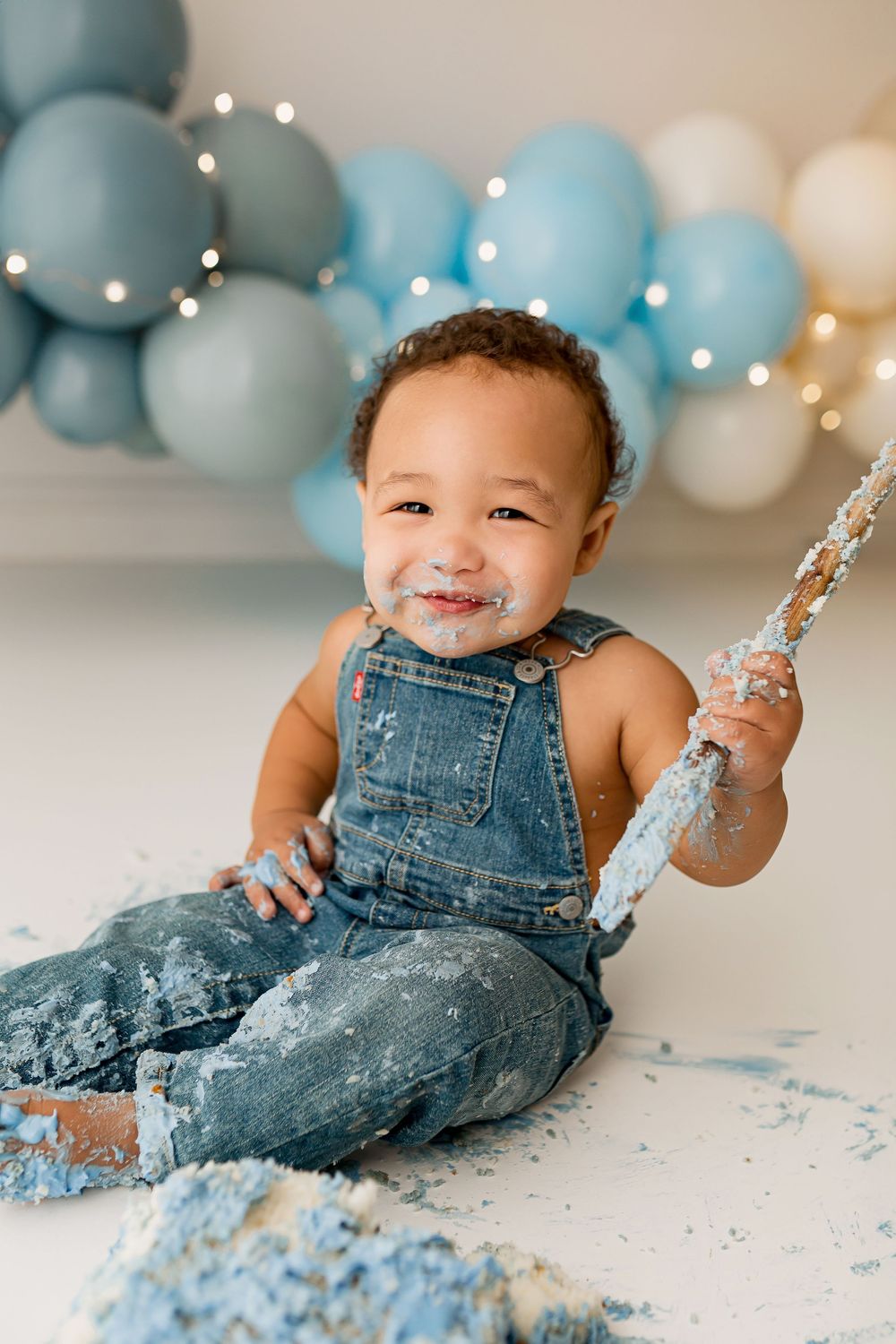 Happy baby in denim overalls enjoying cake smash session with blue balloons at Summerville SC photography studio