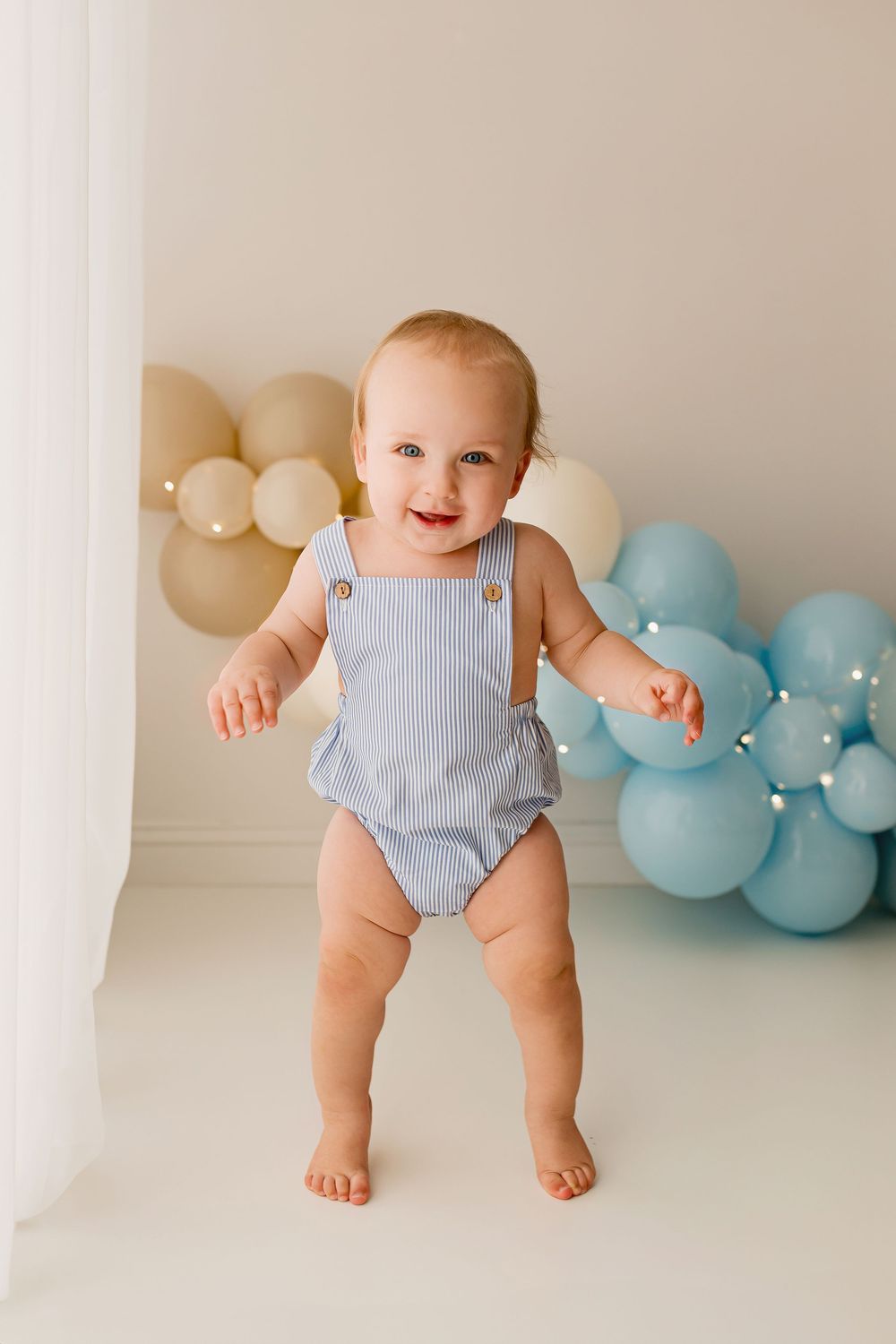 Happy baby in blue striped romper standing with colorful balloon backdrop at Summerville SC photography studio
