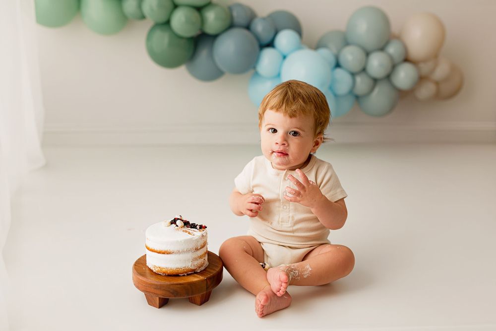 Baby boy in cream outfit sitting next to birthday cake with blue and green balloon backdrop at Summerville SC studio