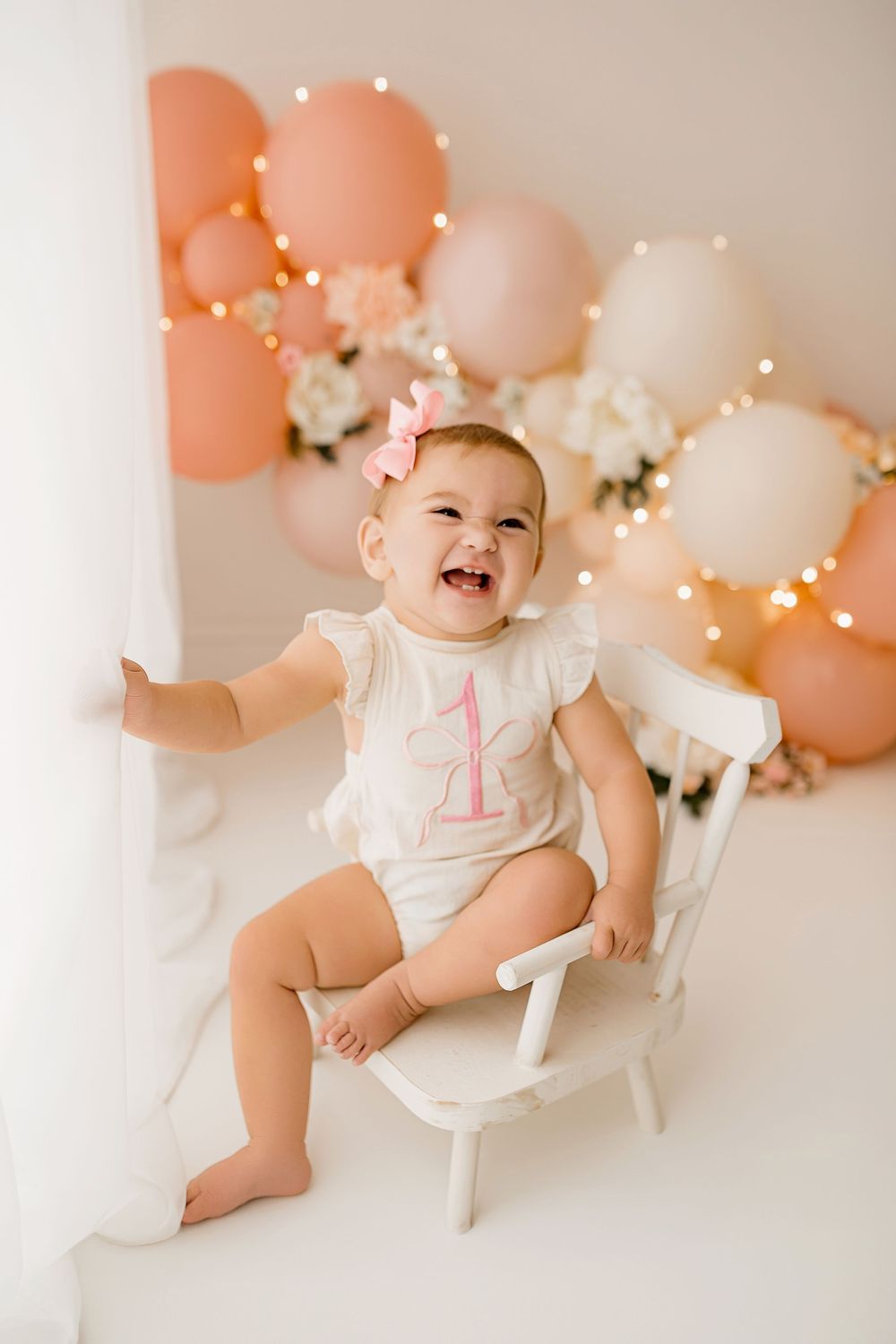 Happy baby girl in white outfit with pink bow celebrating first birthday with balloons in Summerville SC studio