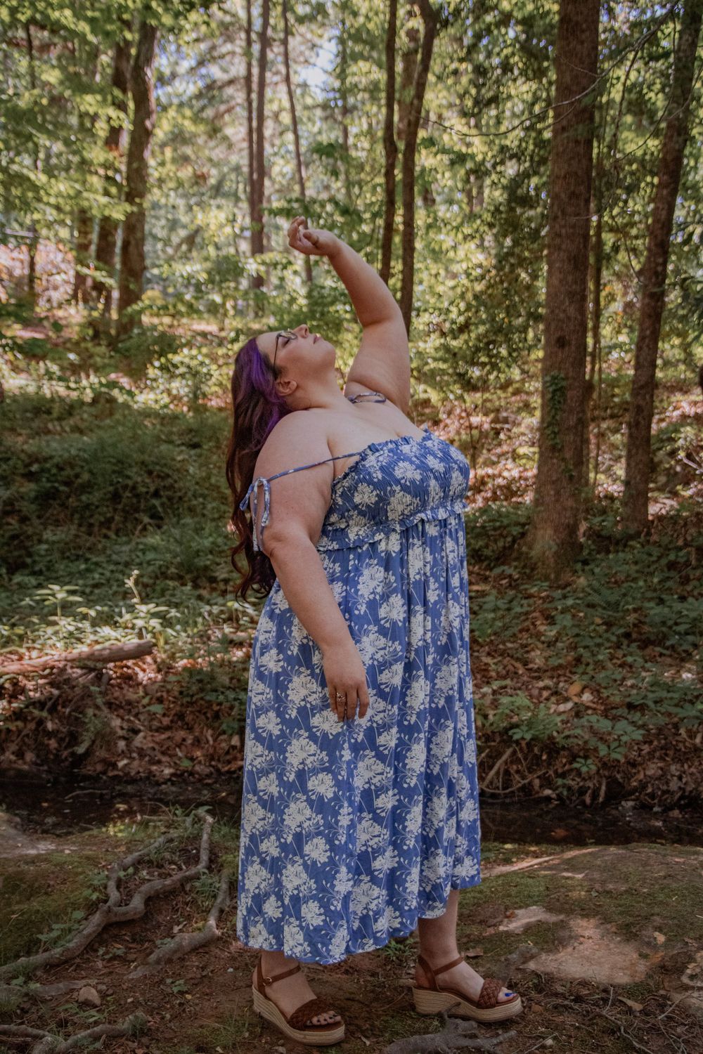 Woman in blue floral maxi dress posing in forest, looking up with arm raised, wearing platform sandals.