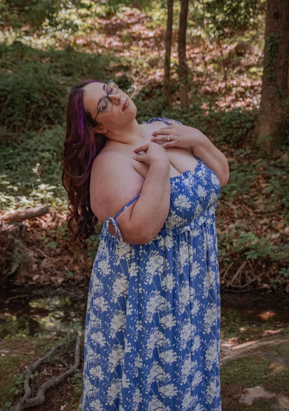 Woman in blue floral sundress posing gracefully in a lush green forest setting with purple hair.