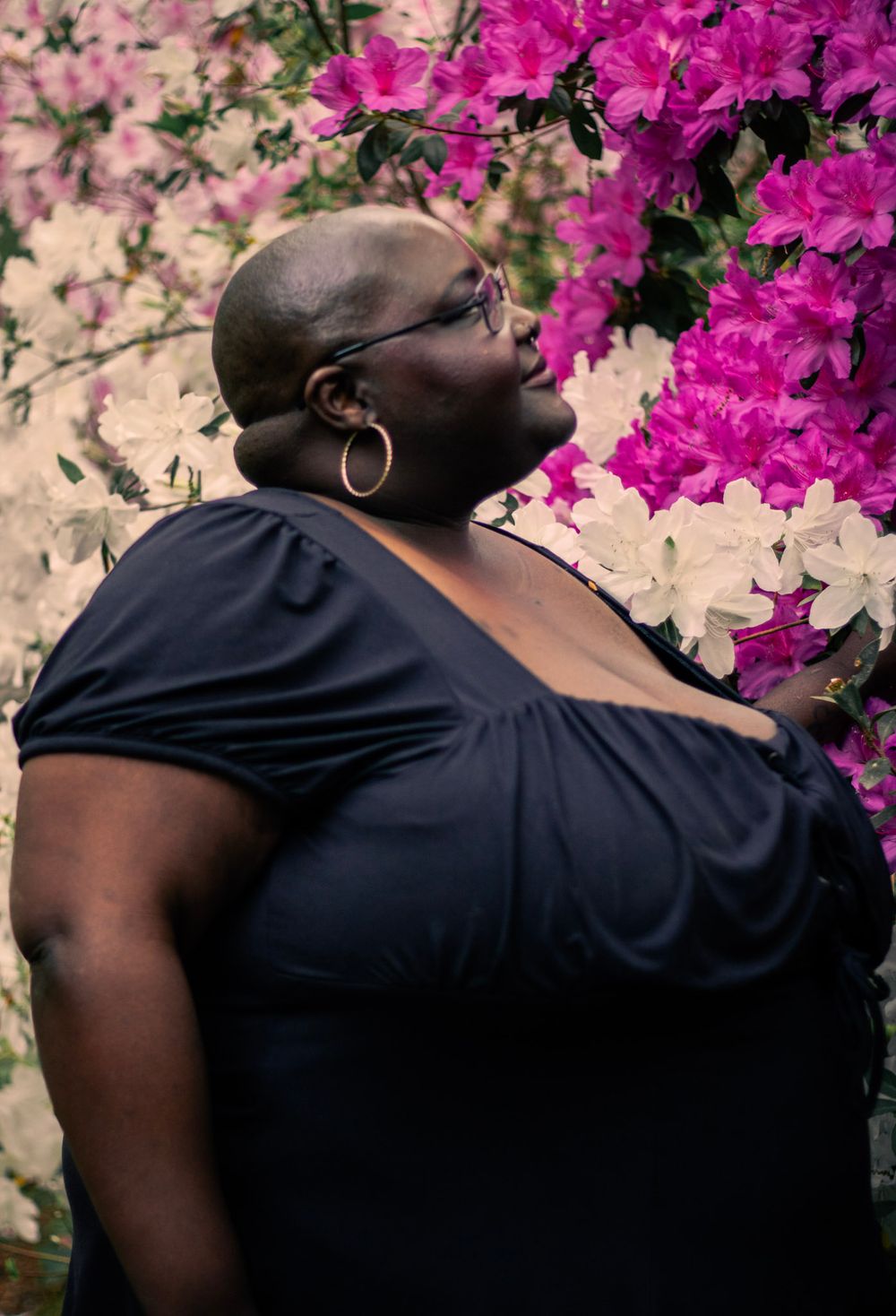 A bald Black woman in a navy top gazes upward beside vibrant pink and white flowering azalea bushes.