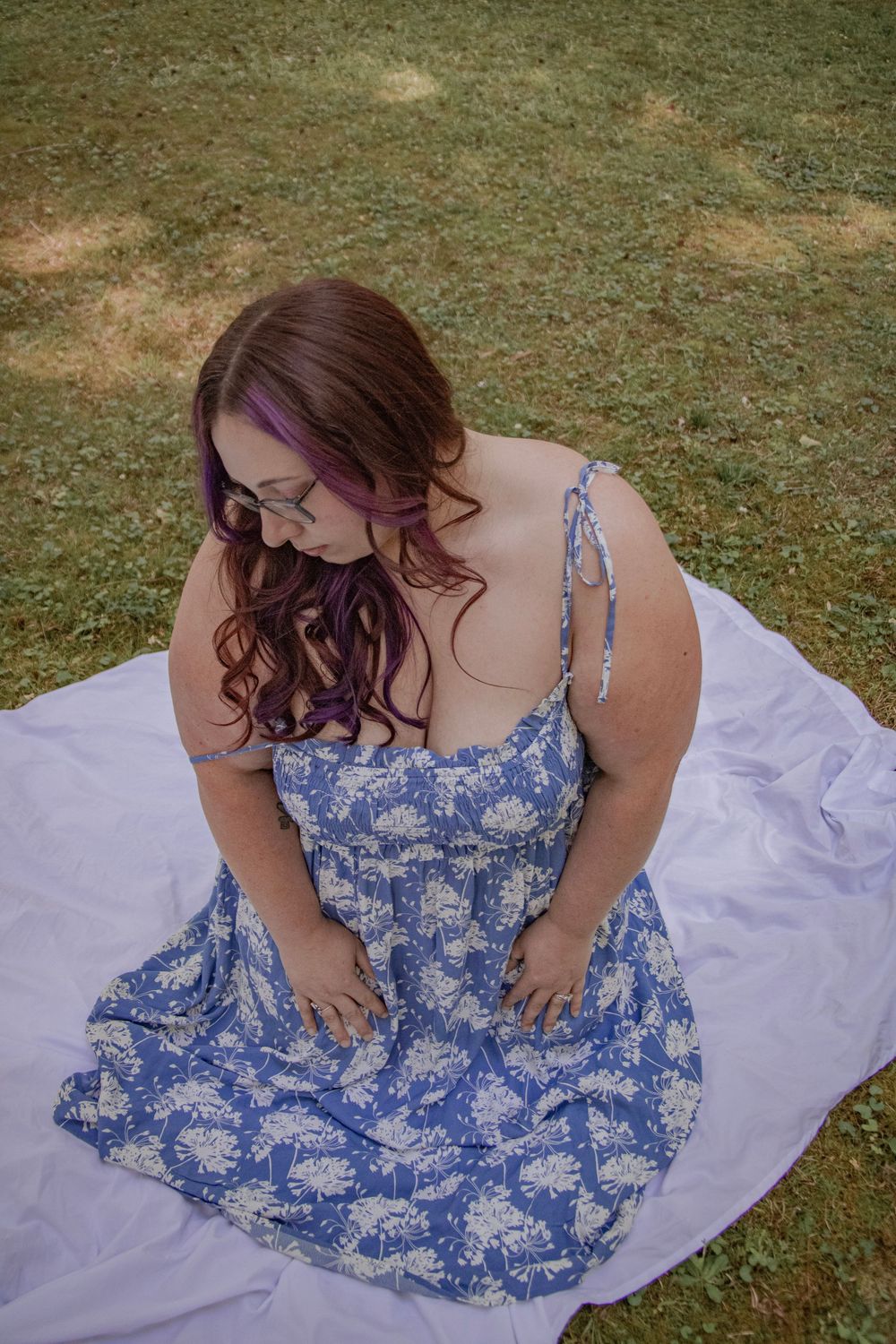 Woman in blue floral sundress sitting on white blanket in grass, looking down, with purple-highlighted hair.