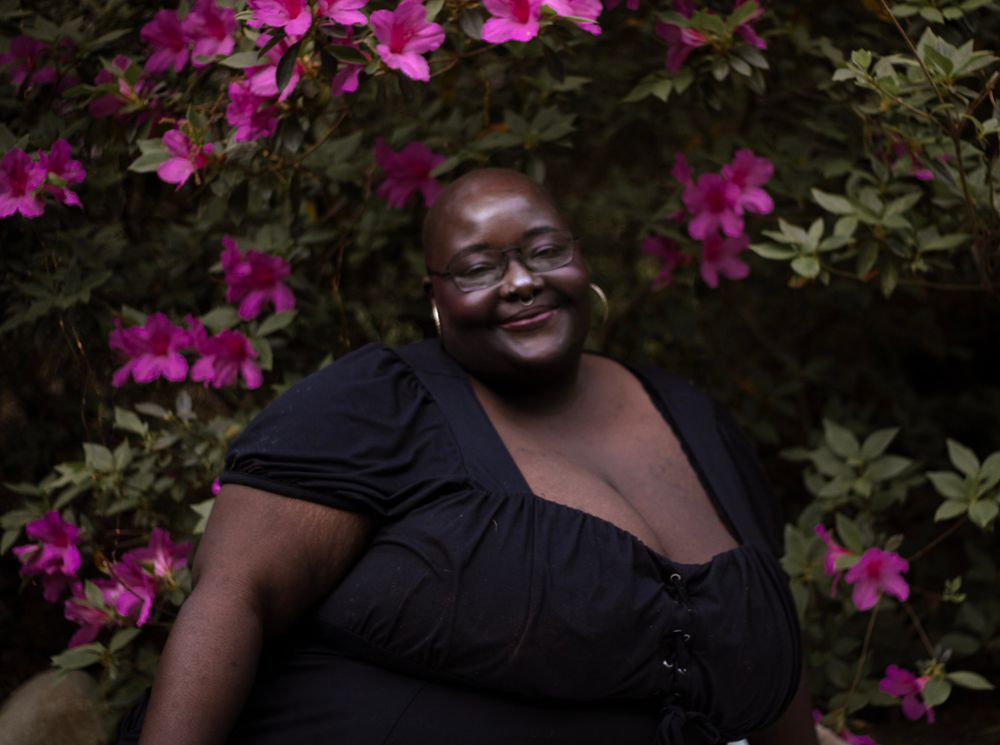 Smiling woman in black top surrounded by vibrant pink azalea flowers in a lush garden setting.