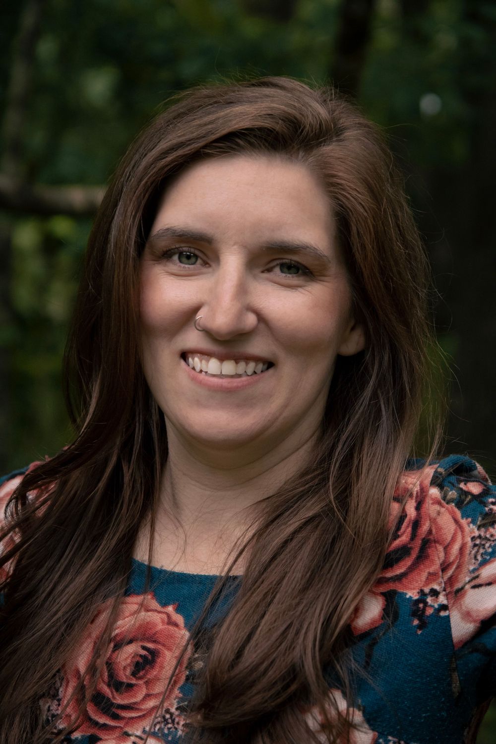 Smiling woman with long brown hair wearing a floral teal dress, photographed outdoors with green foliage background.