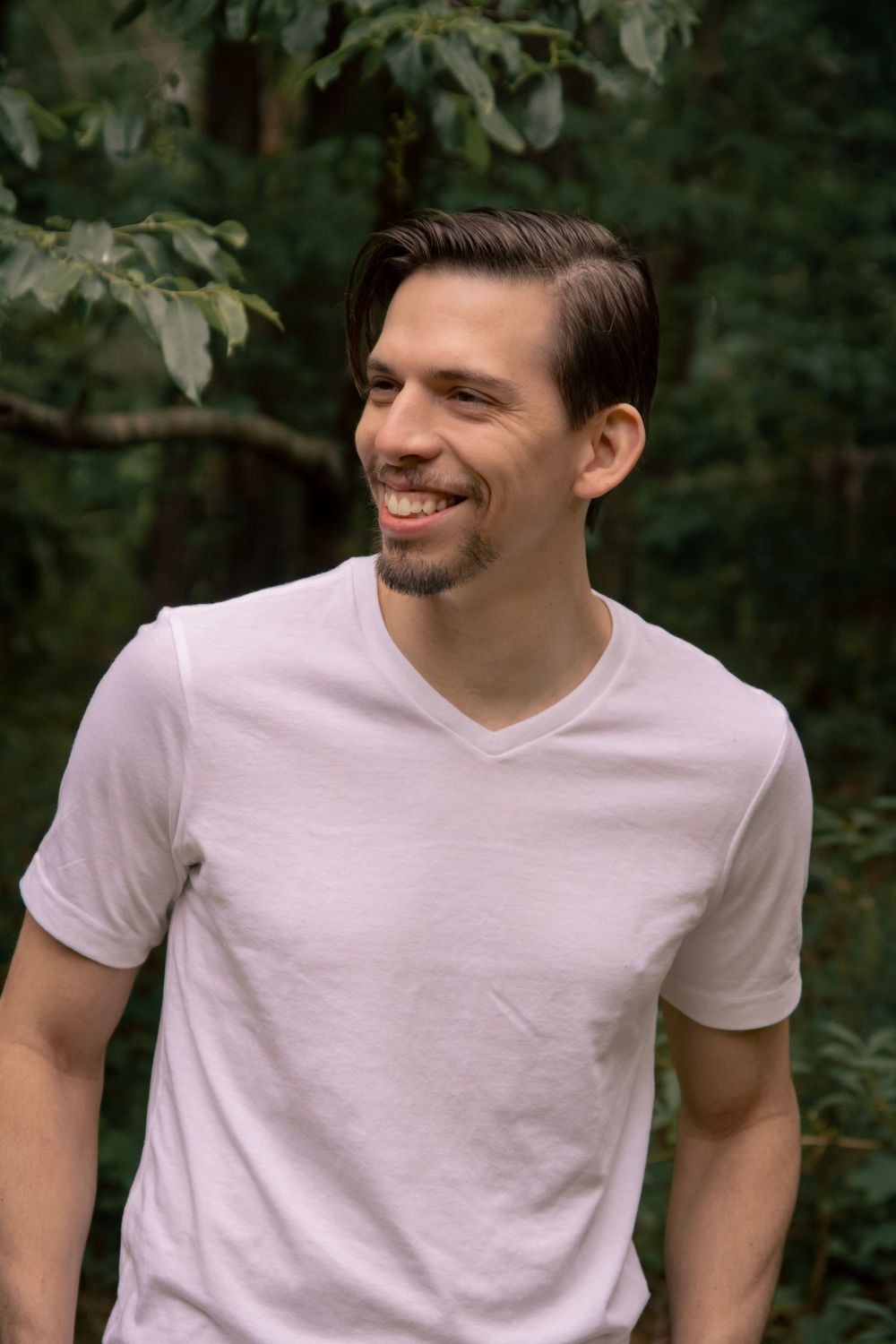 Smiling young man with dark hair wearing a white v-neck t-shirt standing outdoors in a green forest setting.