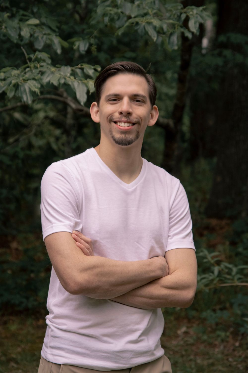 Smiling man with crossed arms wearing a white v-neck t-shirt standing outdoors in front of lush green trees.