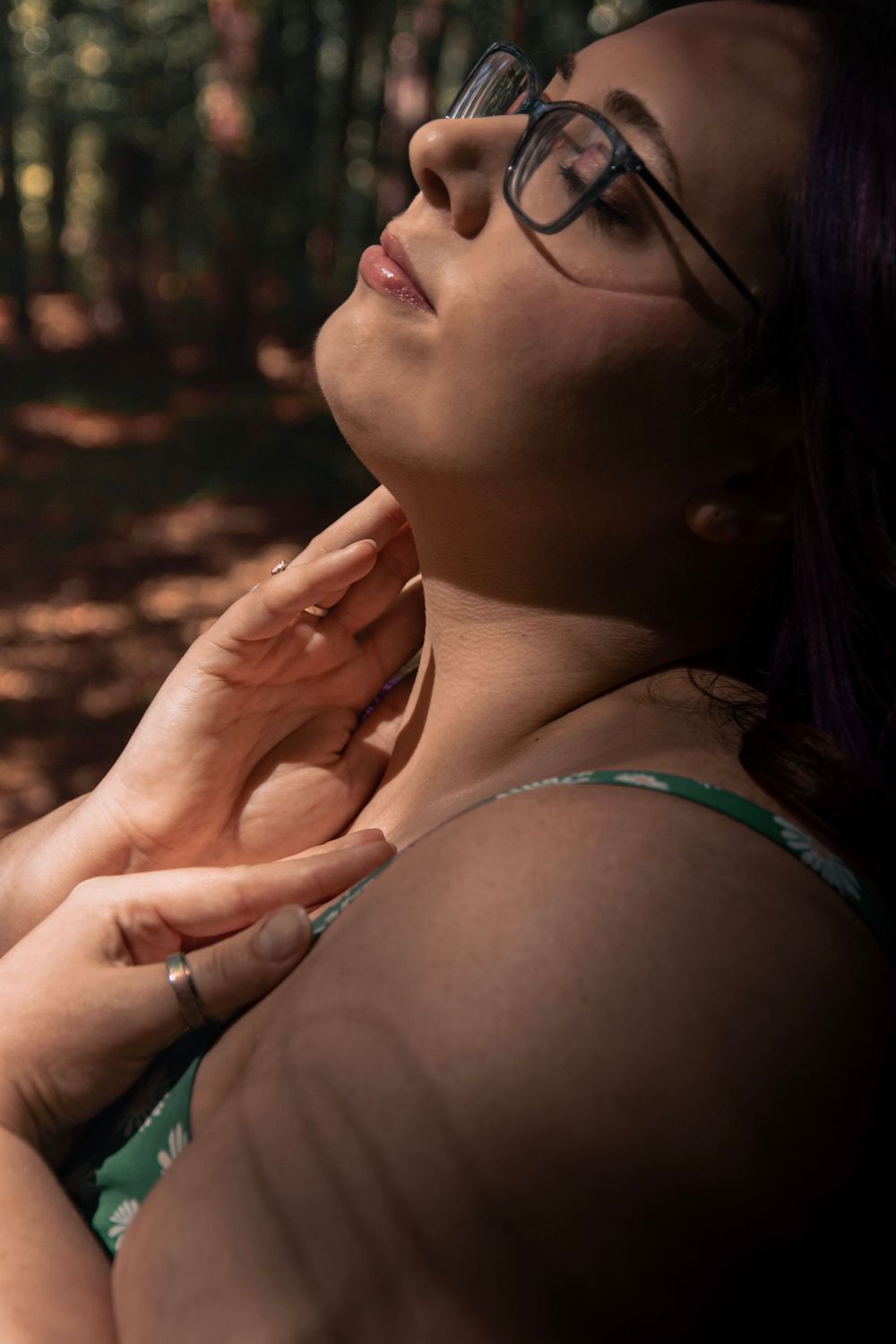 Woman with glasses tilting head back in sunlit forest, hands on neck, wearing green top.