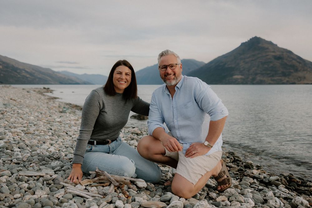 A smiling couple sitting on a rocky lakeshore at Homestead Bay in Queenstown during a family photoshoot at golden hour.