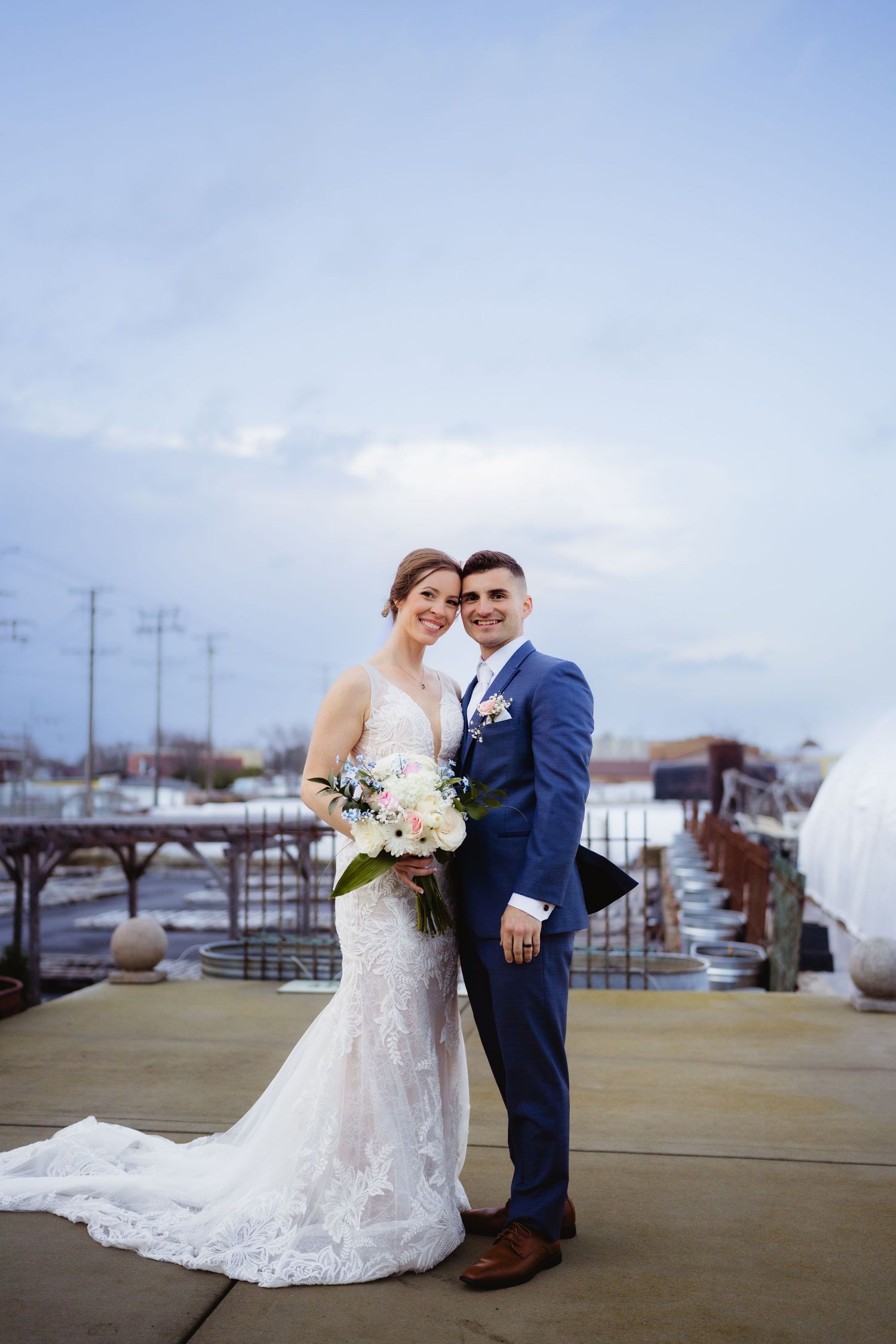 Bride in lace gown and groom in blue suit pose together on rooftop terrace holding floral bouquet on wedding day.