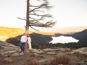 A couple stands together near a barren tree on a rocky overlook with a lake and mountains in the golden sunset.