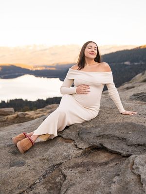 A woman in an off-shoulder dress sits on rocky terrain during golden hour with a lake view.