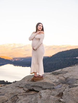 A person in a cream dress poses on a rocky cliff overlooking a serene lake at sunset in a mountain landscape.