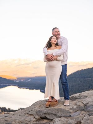 A couple embraces on a rocky cliff overlooking a scenic mountain lake during sunset.