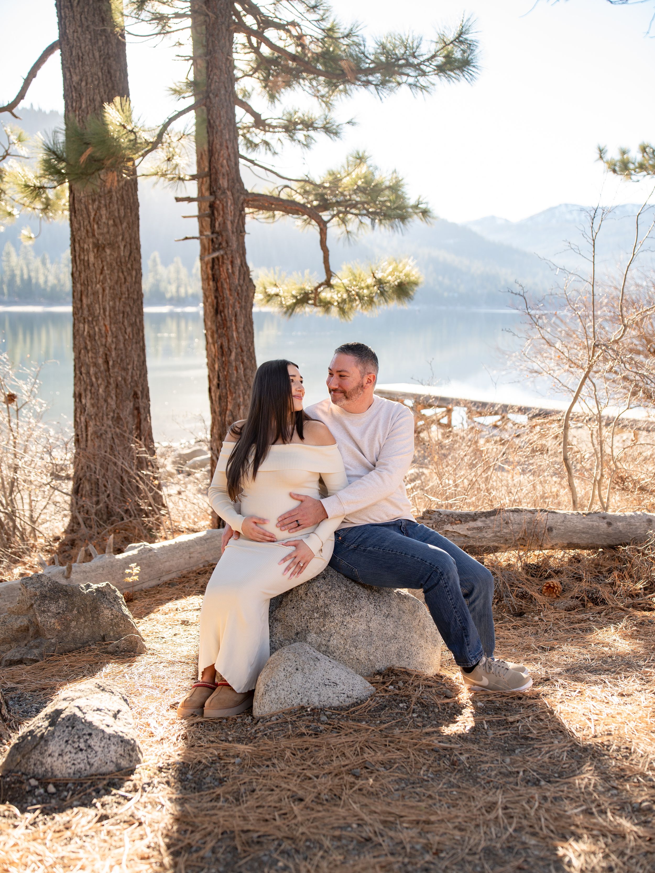 A couple sits together on a large rock near a scenic mountain lake on a sunny day.
