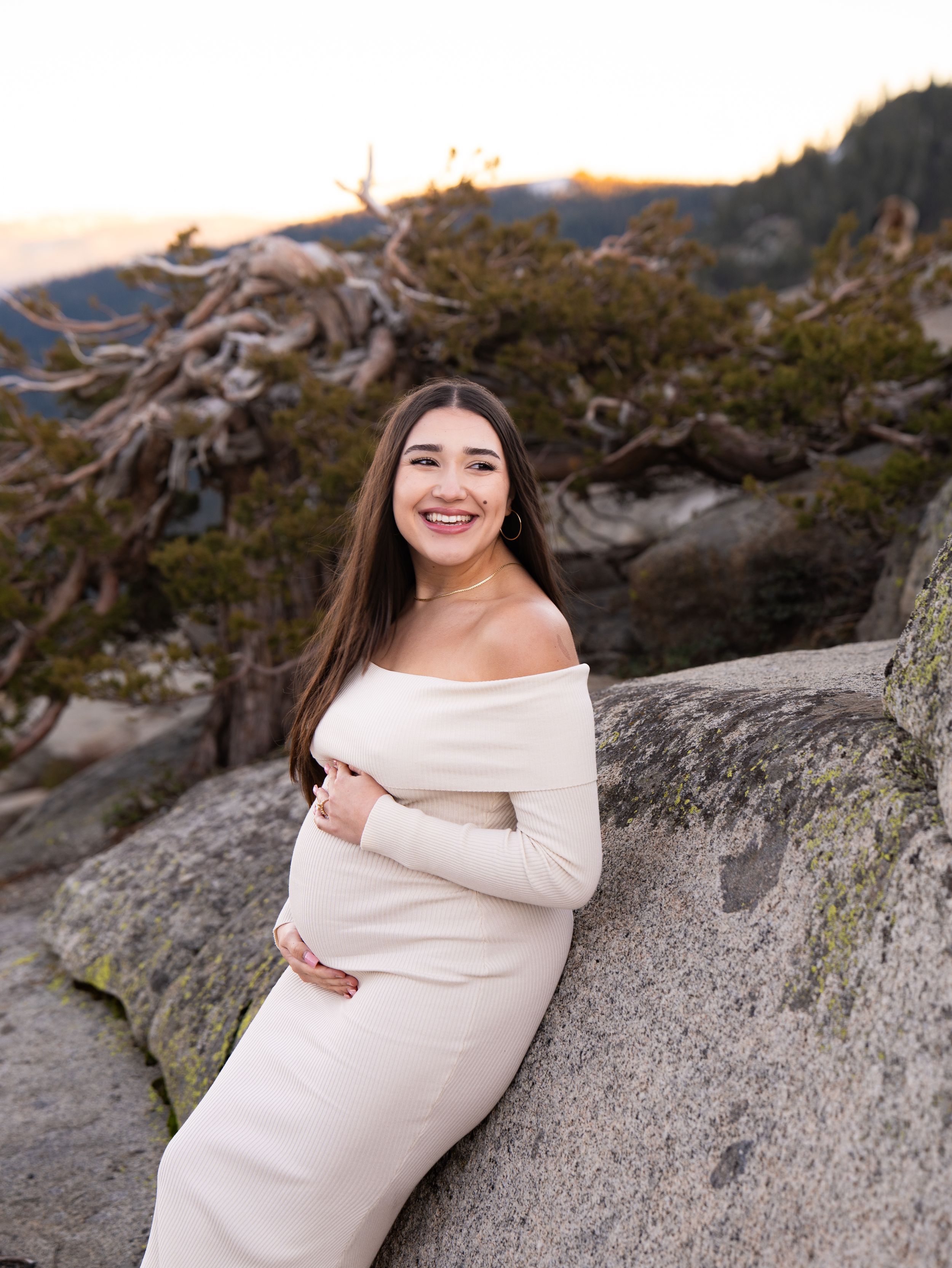 An expectant mother in a cream dress poses on rocky terrain during sunset.