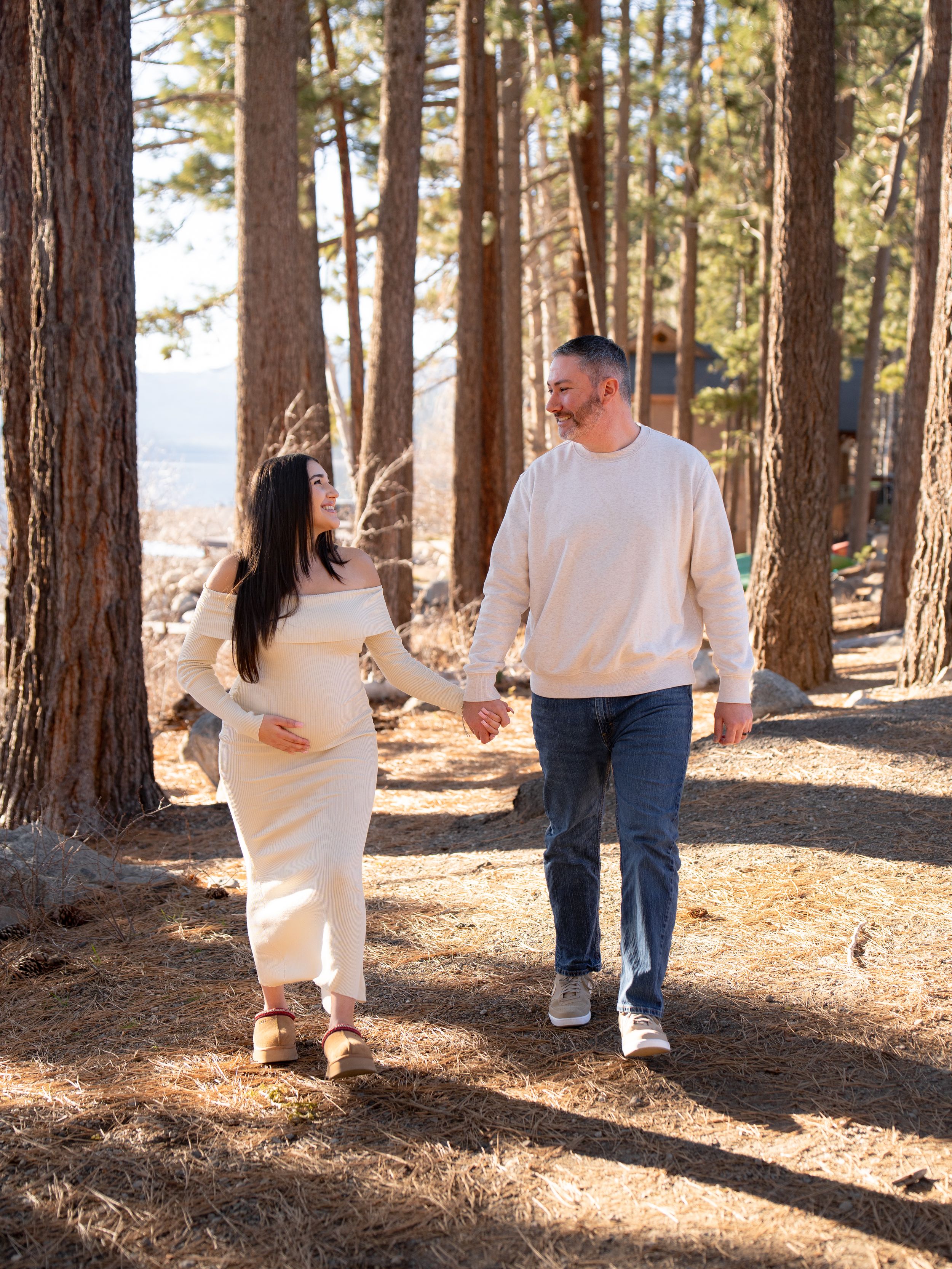 A couple walks hand in hand along a sunlit forest path surrounded by tall pine trees.