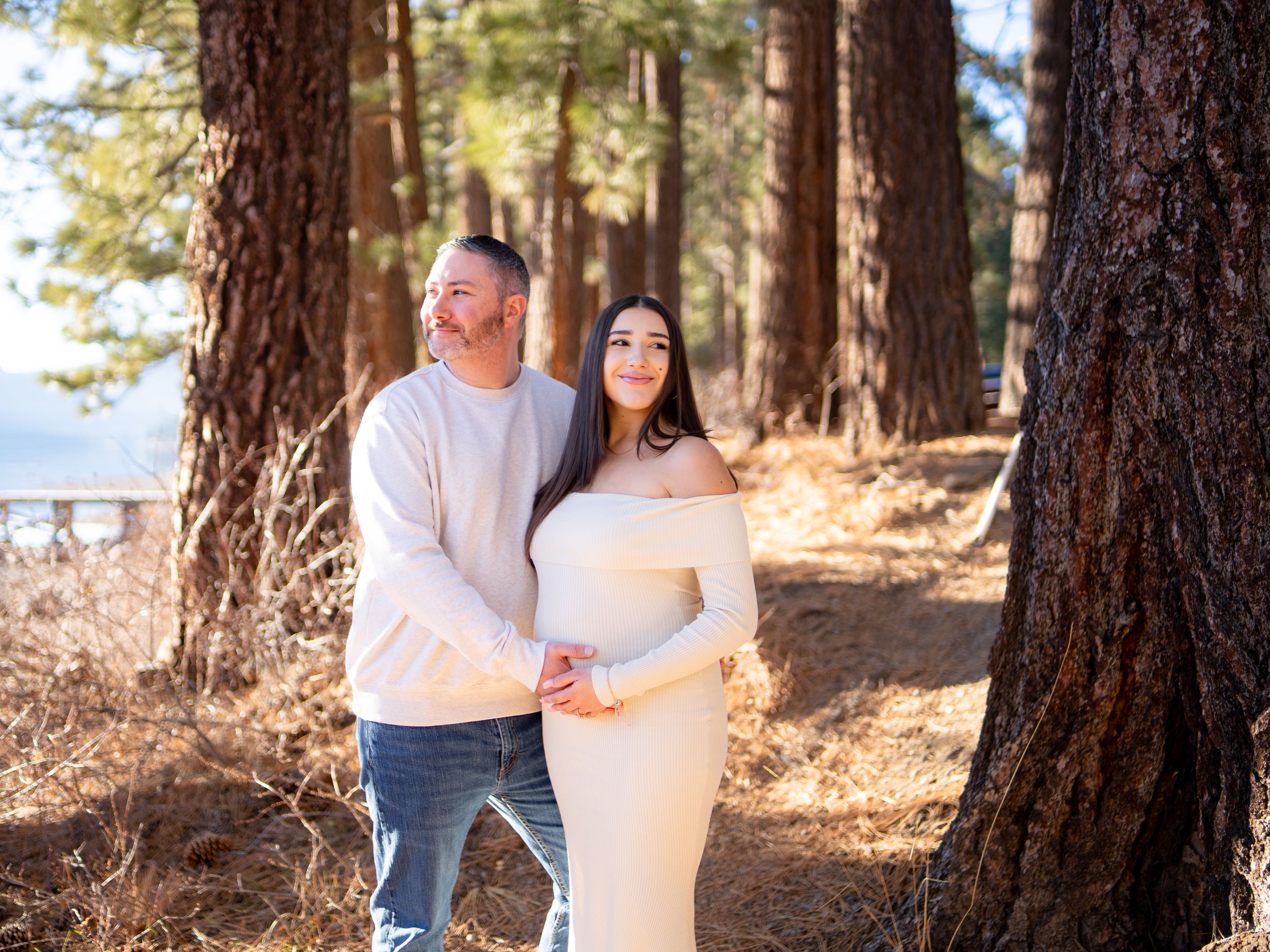 A couple embraces while walking among tall pine trees in a sunlit forest during an outdoor engagement photoshoot.