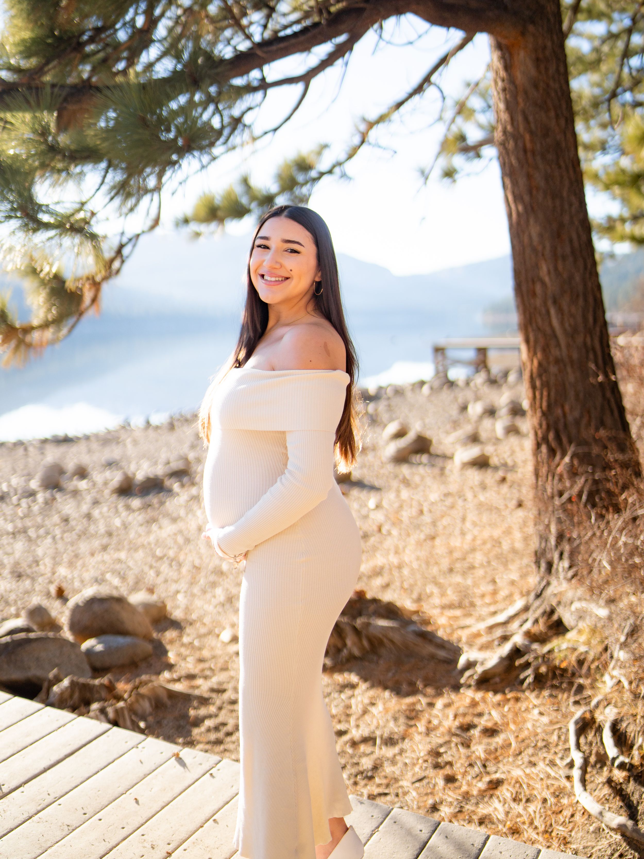 Maternity photoshoot in a scenic mountain forest setting with sunlight filtering through pine trees.