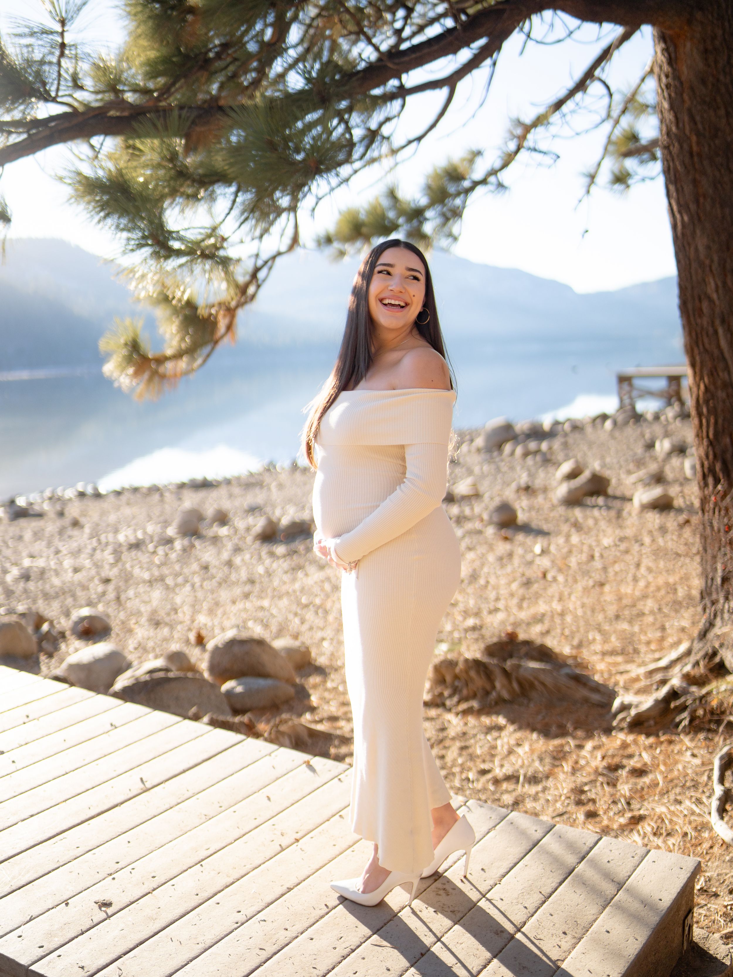A woman in a white dress poses on a wooden deck overlooking a scenic mountain lake.