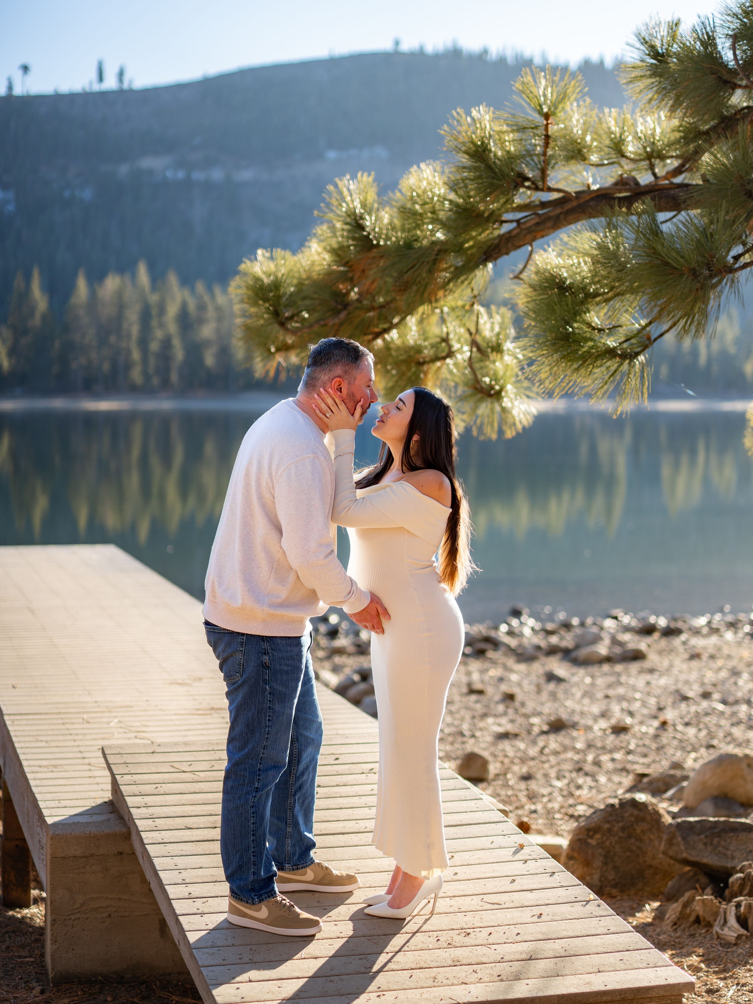 A romantic moment unfolds on a wooden dock overlooking a serene mountain lake surrounded by pine trees.