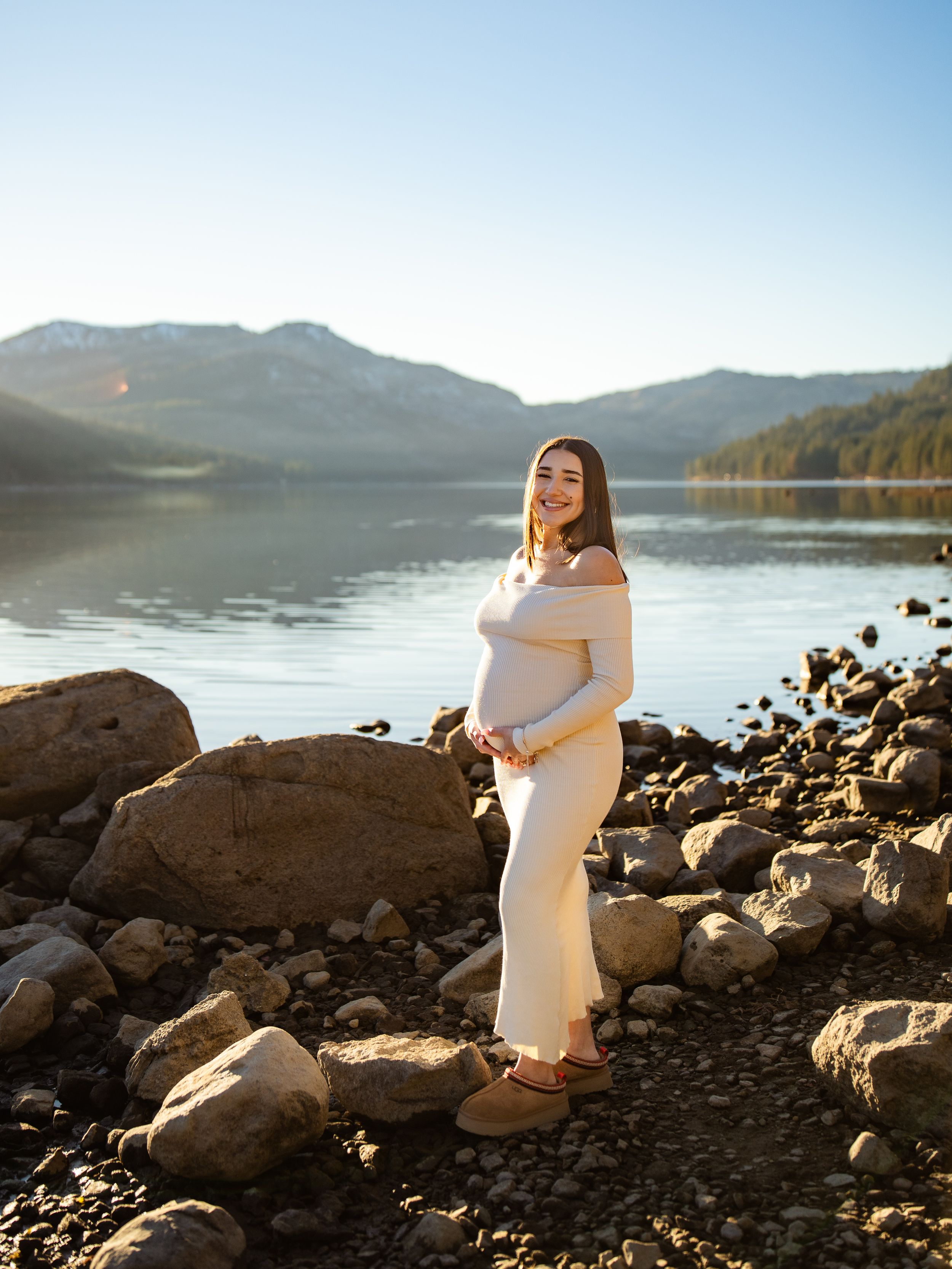 Maternity photos captured on a rocky lakeshore with mountains and calm waters in the background.
